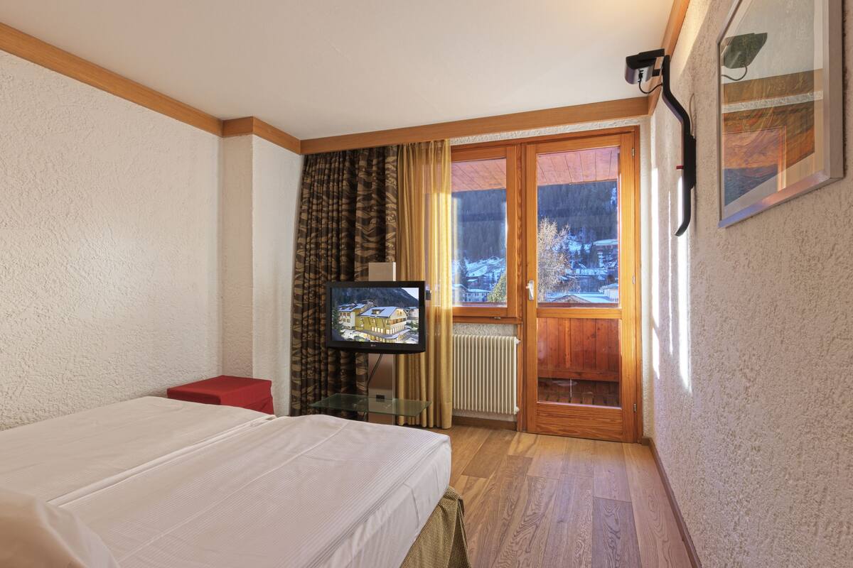 Una camera d'albergo con un letto, una televisione, un tavolo rosso, una finestra vista su un paesaggio innevato e una porta che dà su un balcone.