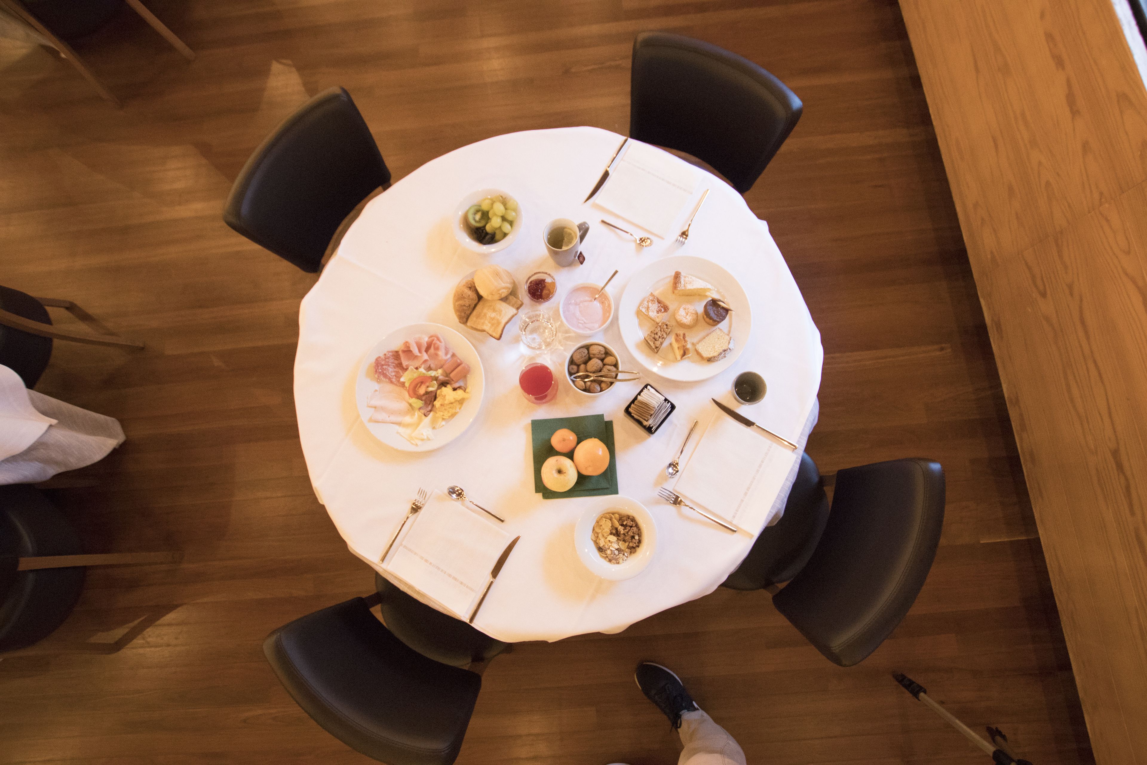 Mesa redonda com diversos alimentos e utensílios, rodeada por cadeiras.