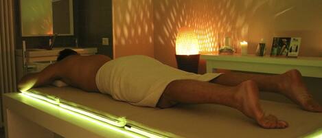 A person lying on a massage table in a room with a mirror, a shelf with candles, and a wall with patterned shadows.