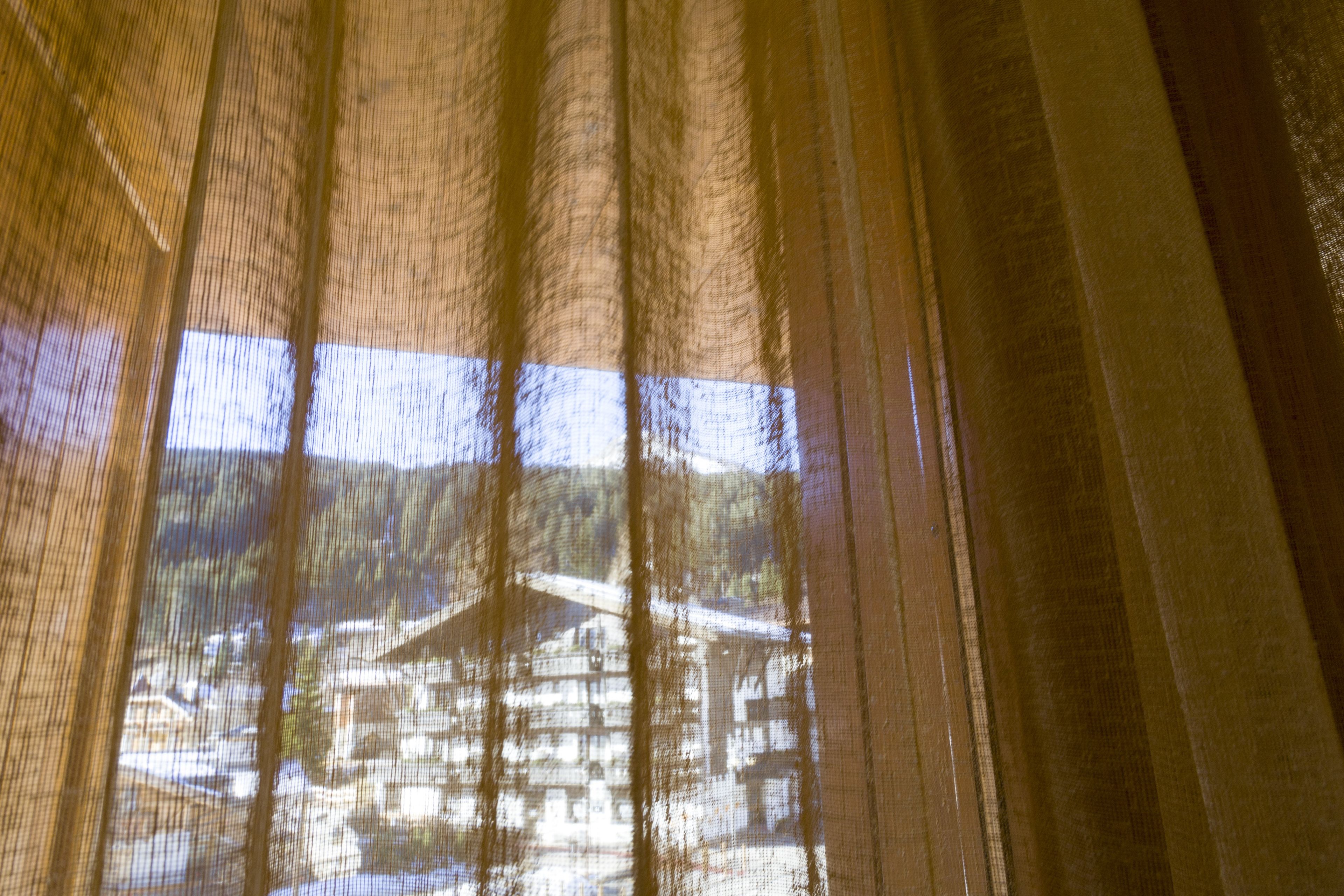 A view through sheer curtains, revealing a building with multiple windows and a roof structure.