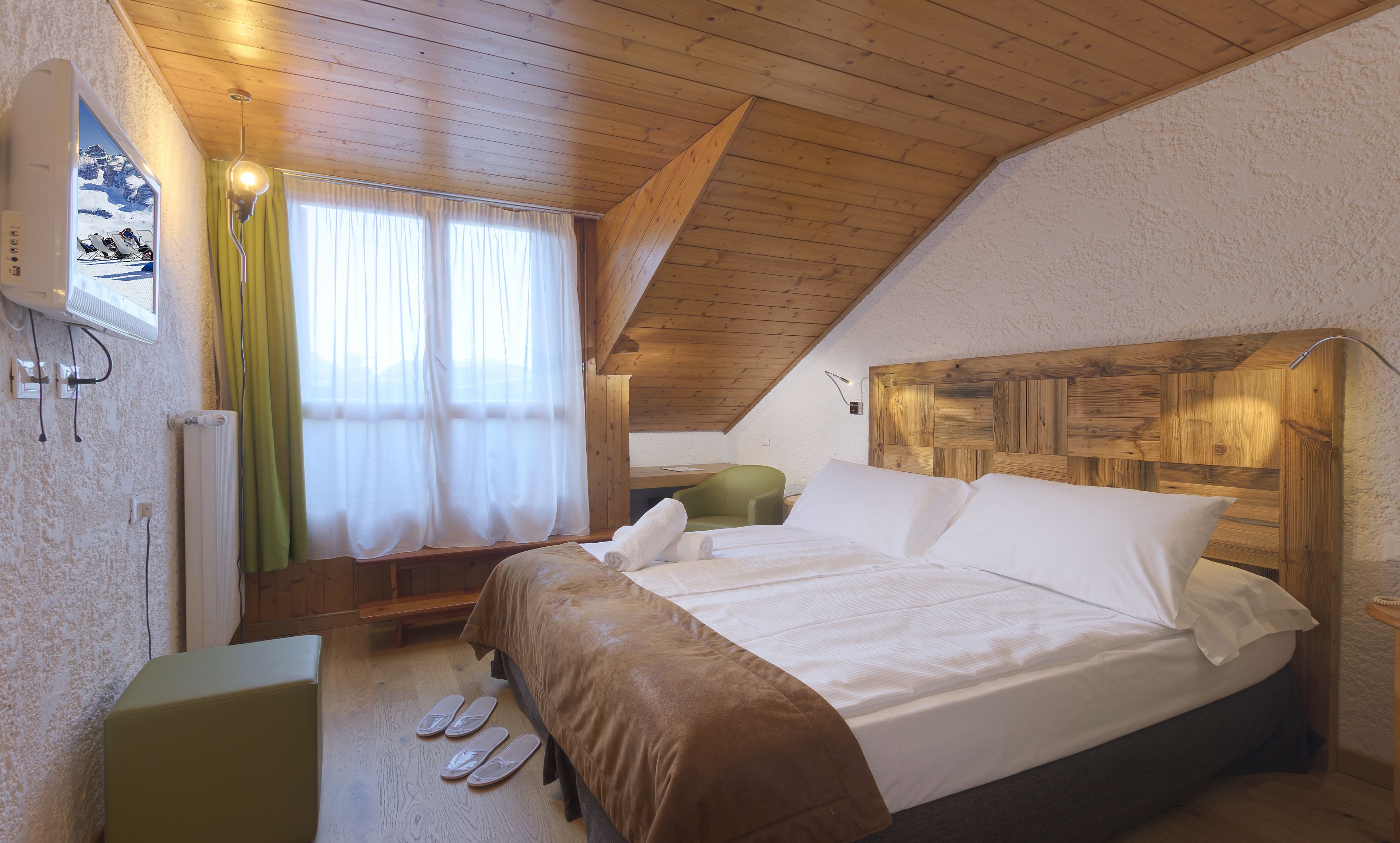 A bedroom with a wooden ceiling, a bed with white linens, a green chair, and a window with sheer curtains.