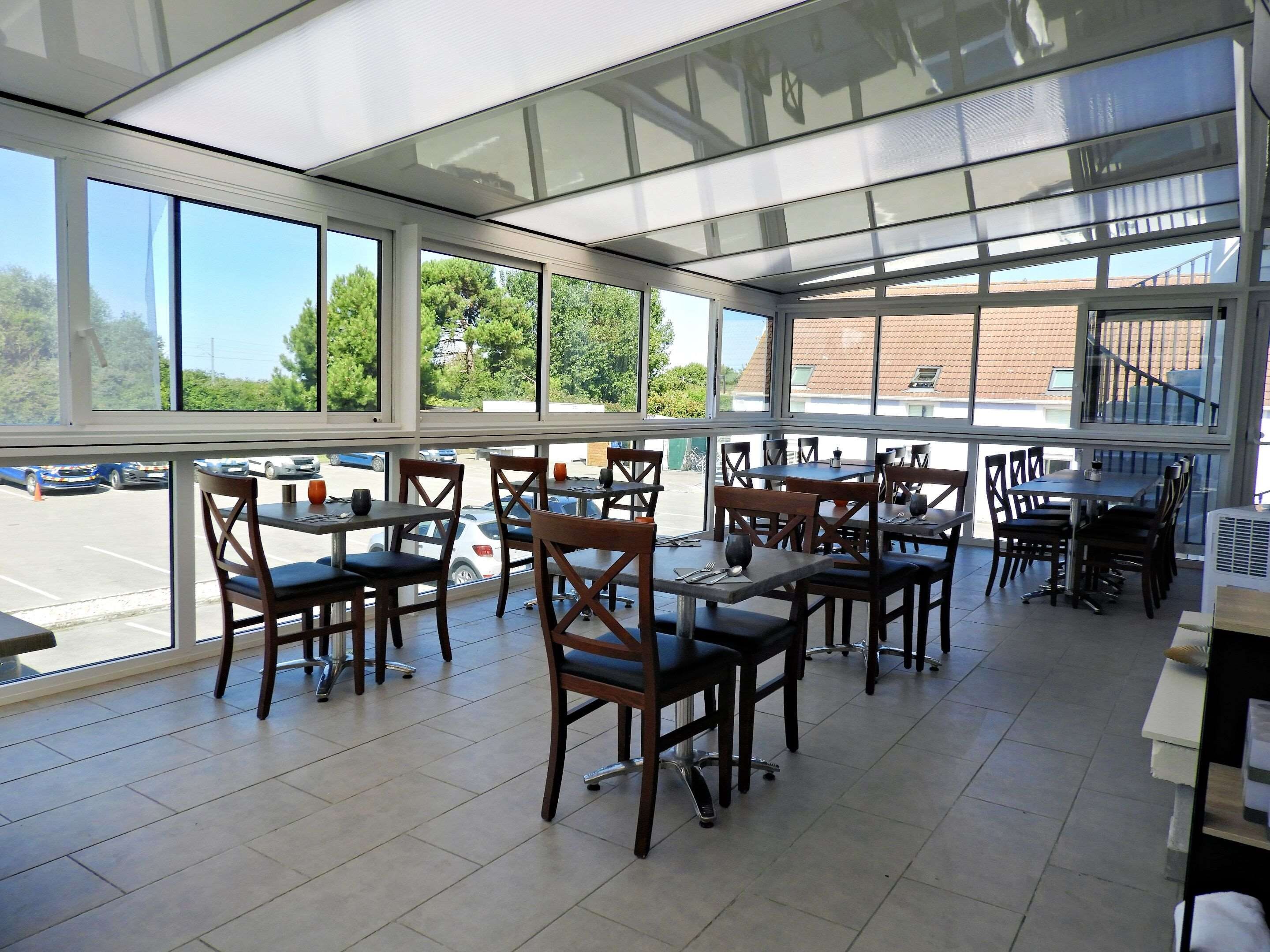 An indoor dining area with multiple tables and chairs, large windows offering a view of a parking lot and greenery outside.