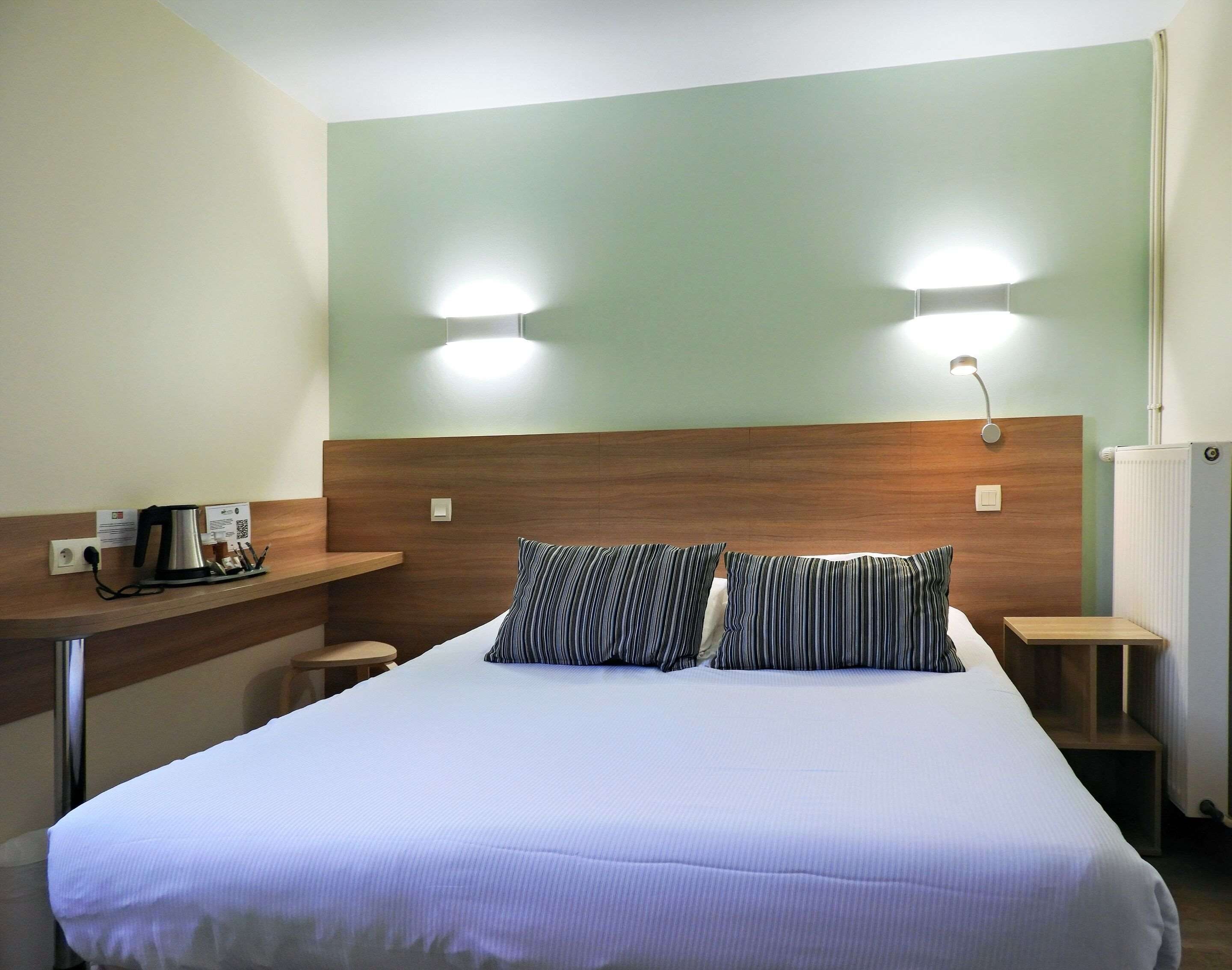Habitación de hotel con cama, dos lámparas de noche, cabecera de madera, una mesita con hervidor y un radiador.