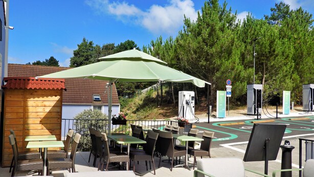 An outdoor seating area with tables and chairs, a green umbrella, and electric vehicle charging stations. - Brit Hotel Le 940 Le Touquet – Etaples (Etaples)