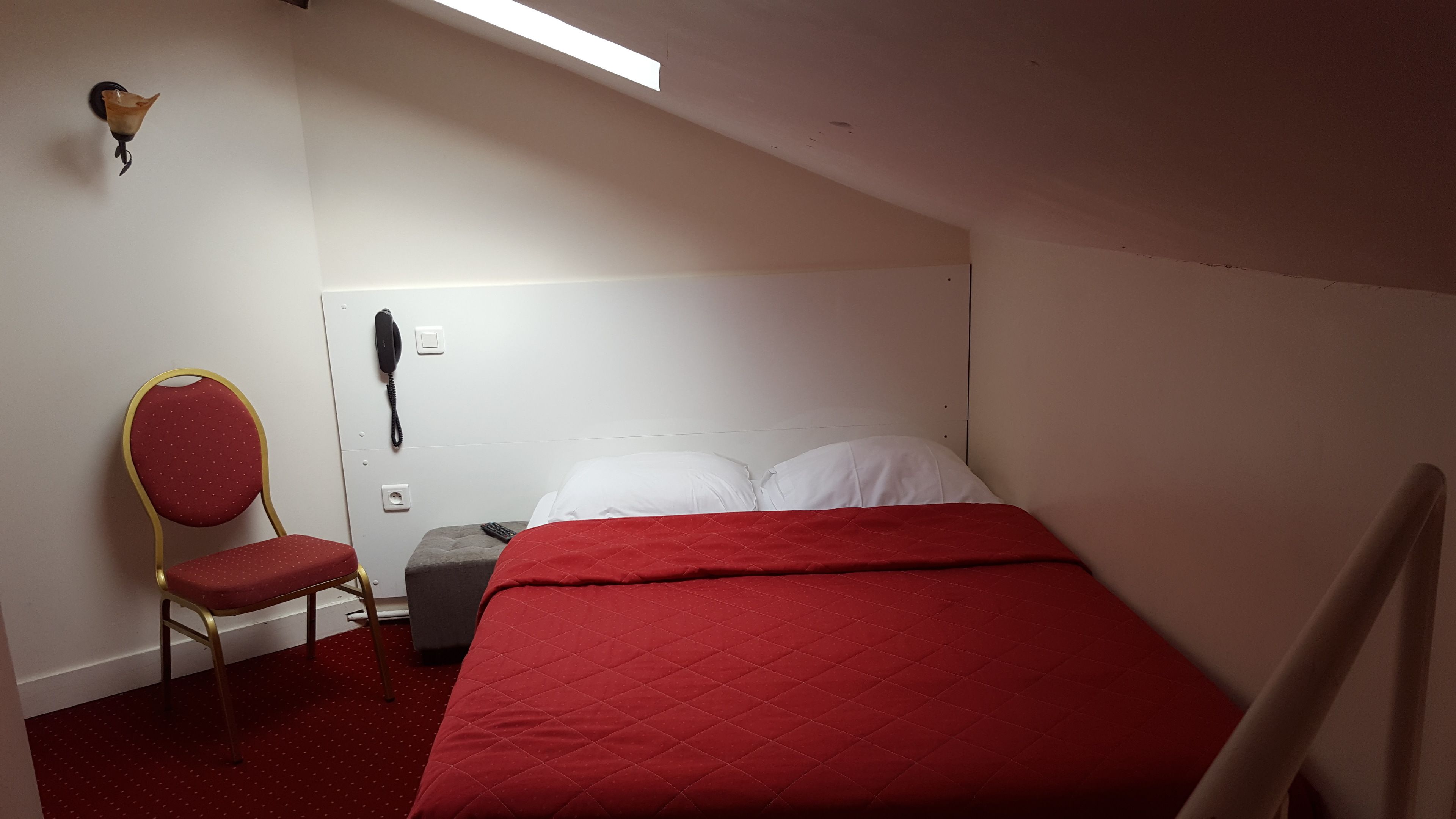 A small, simple room with a bed, a chair, a bedside table, and a phone.
