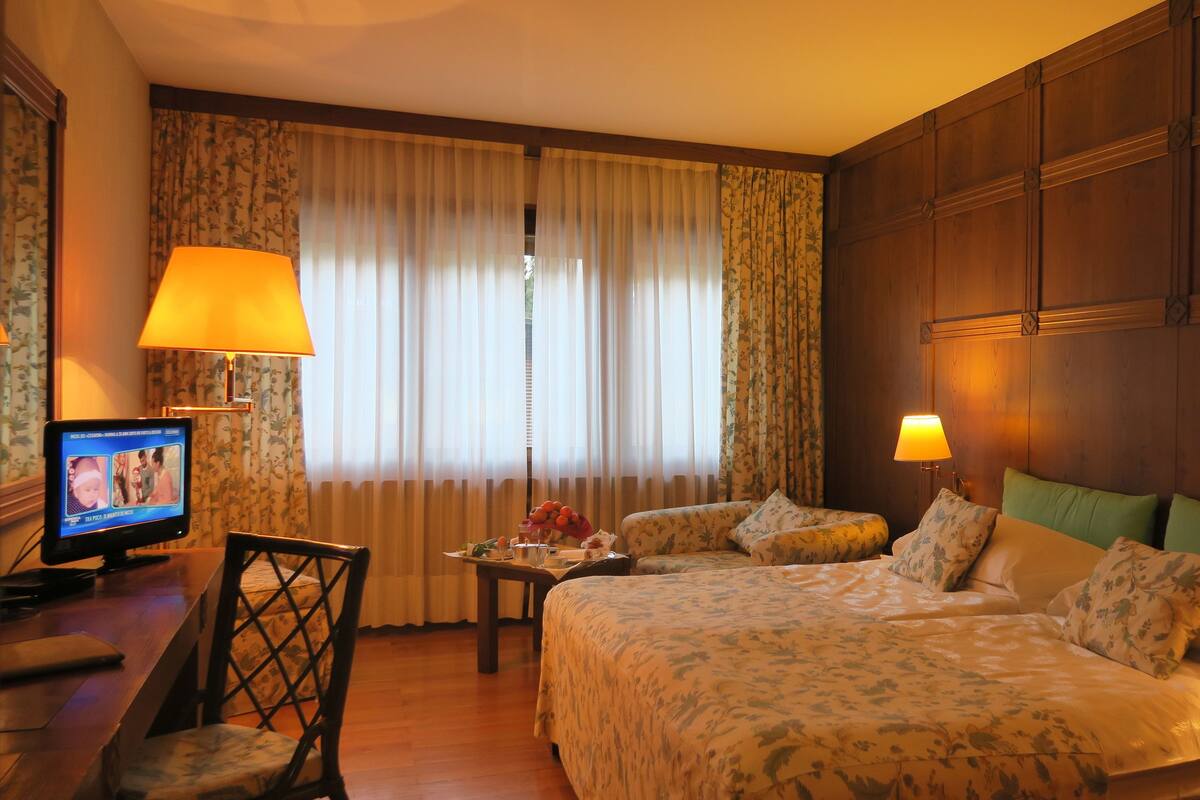 A hotel room with a bed, a desk with a computer, a chair, and a window with curtains.