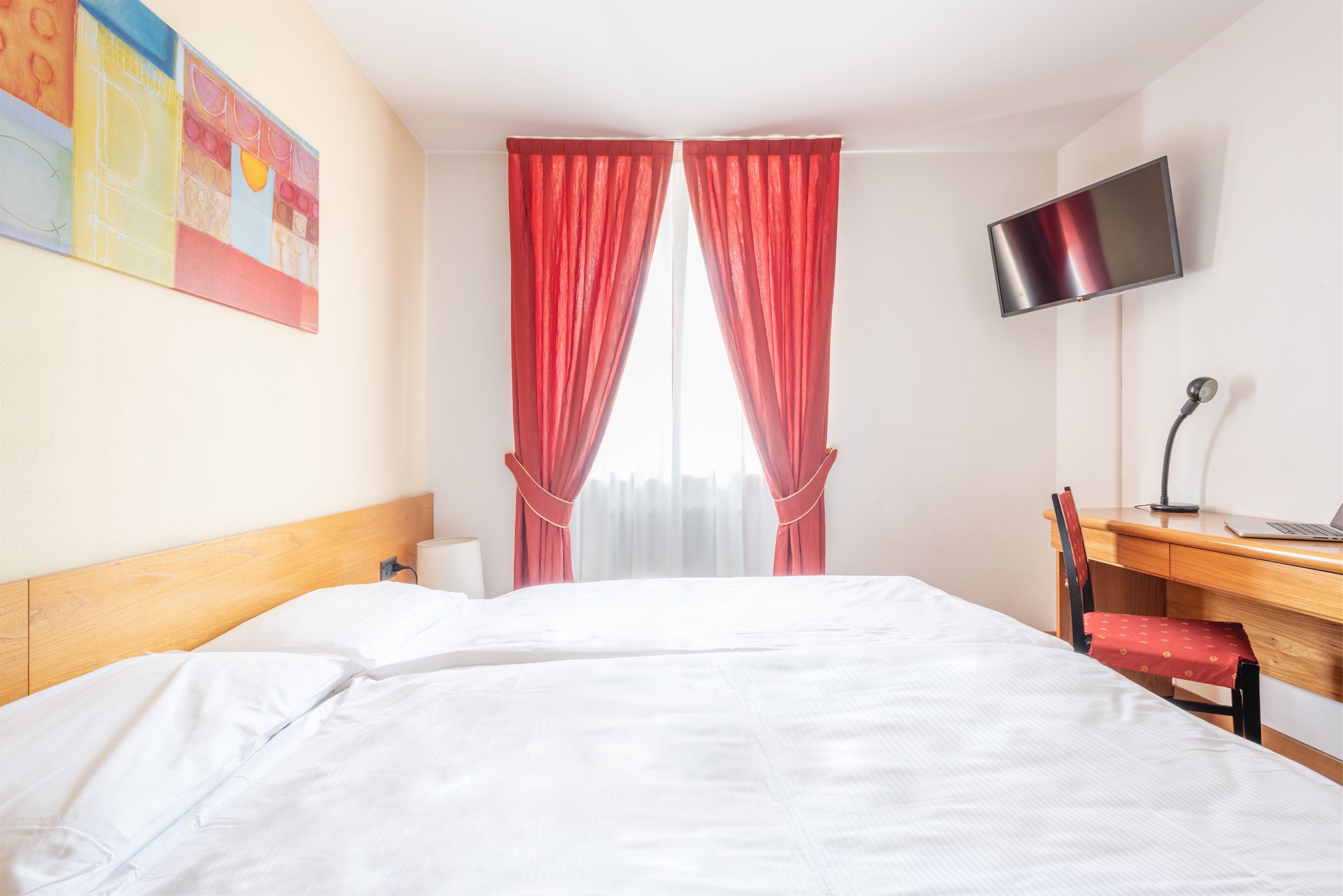 A hotel room with a bed, a desk, a chair, a TV, and a window with red curtains.