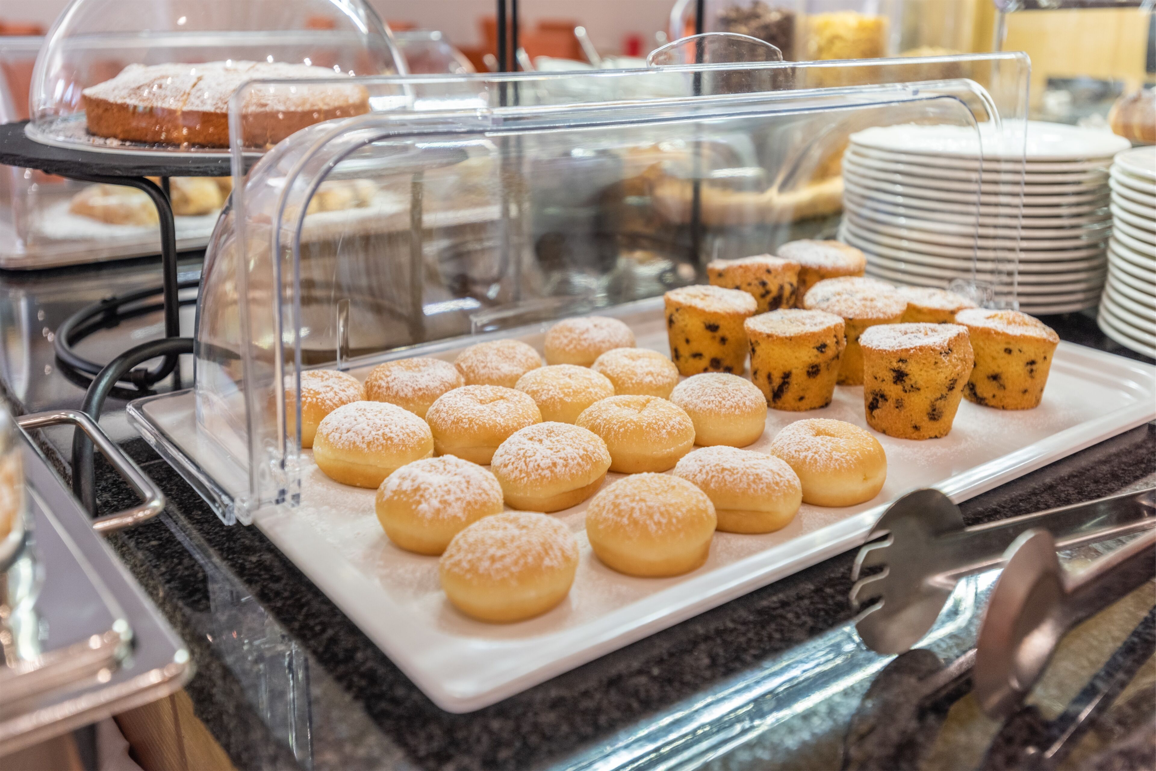 Un bufé con diversos pasteles, entre ellos donuts y muffins, exhibidos en recipientes transparentes.