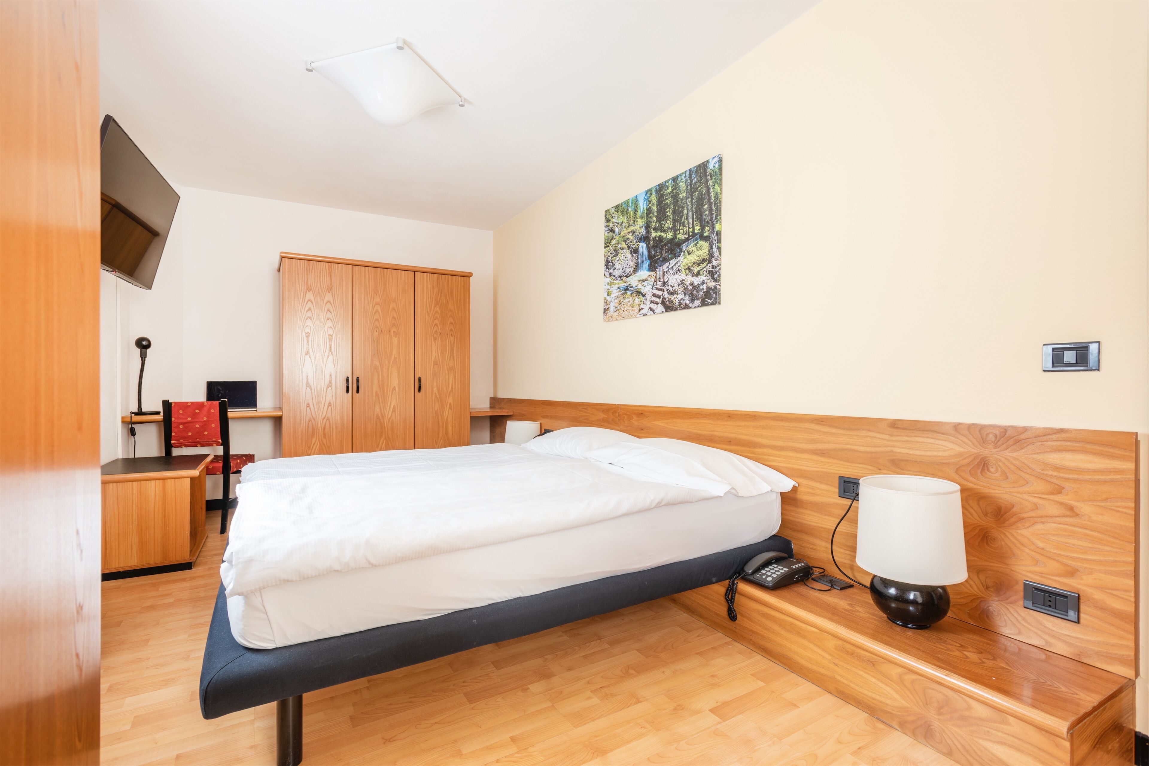 A hotel room with a bed, wooden wardrobe, desk, chair, lamp, and a painting on the wall.