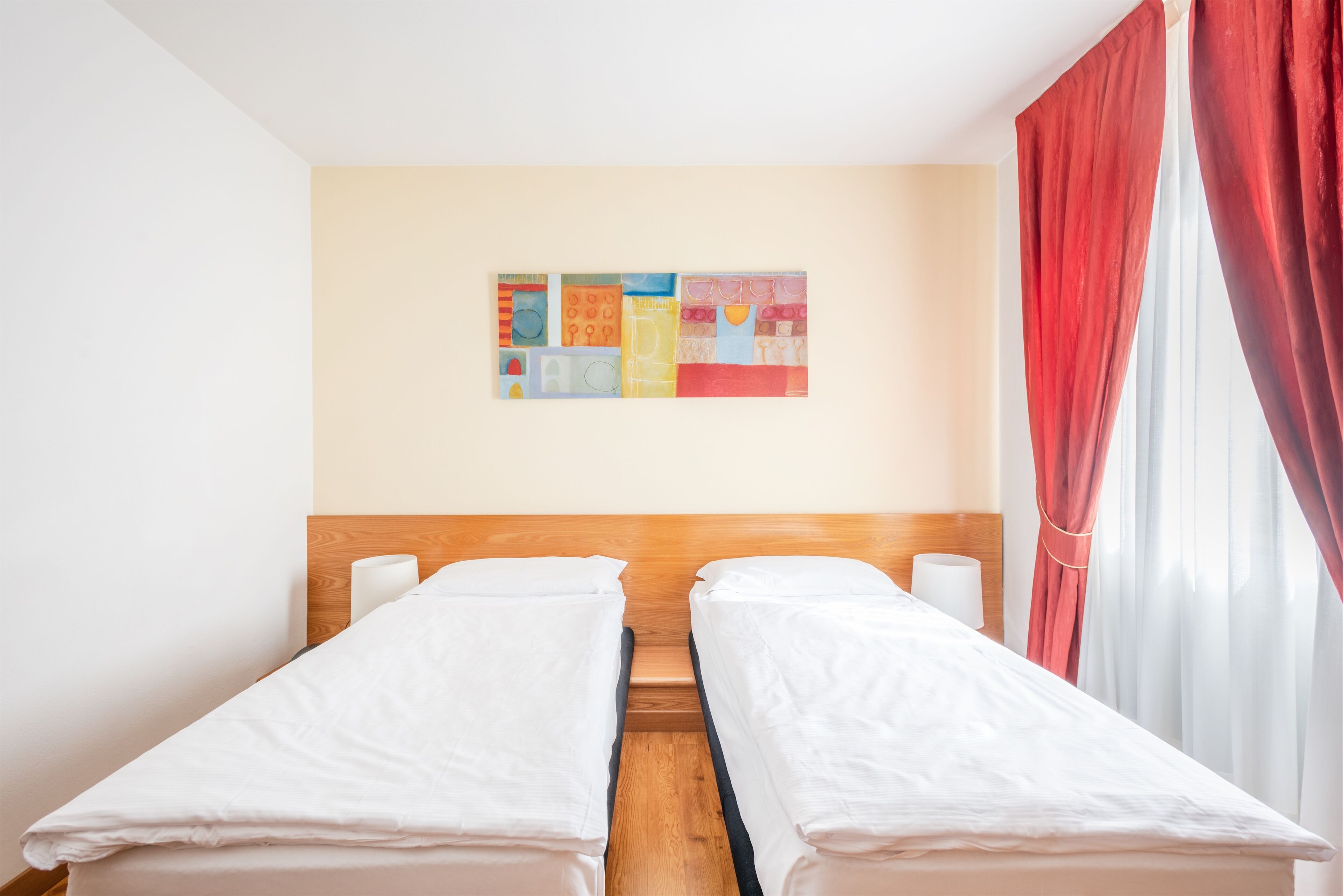 Ein Hotelzimmer mit zwei Einzelbetten, einem Holzkopfende, einem Wandbild und roten Vorhängen.