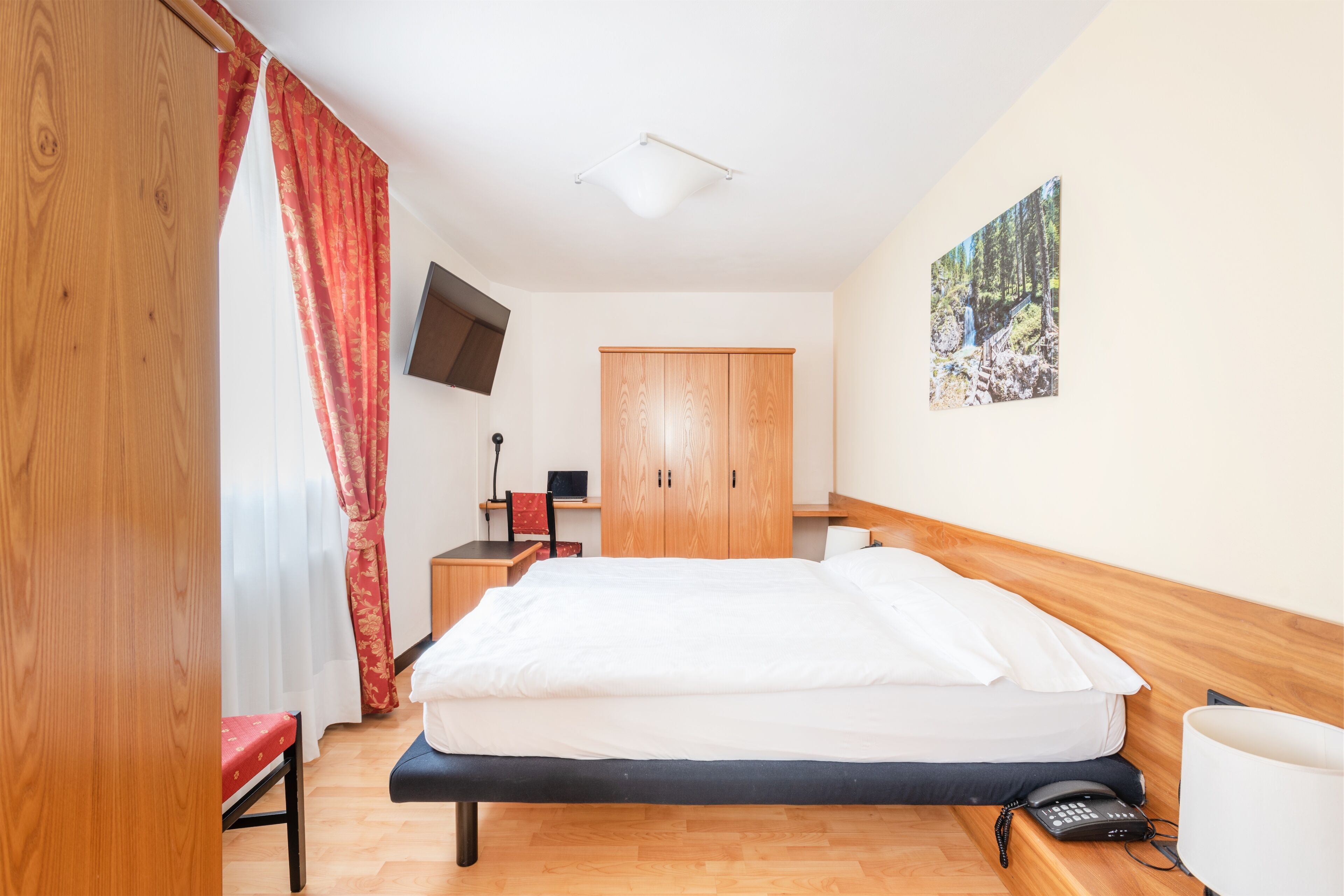 A hotel room with a bed, a desk, a chair, a wardrobe, a TV mounted on the wall, and a painting.