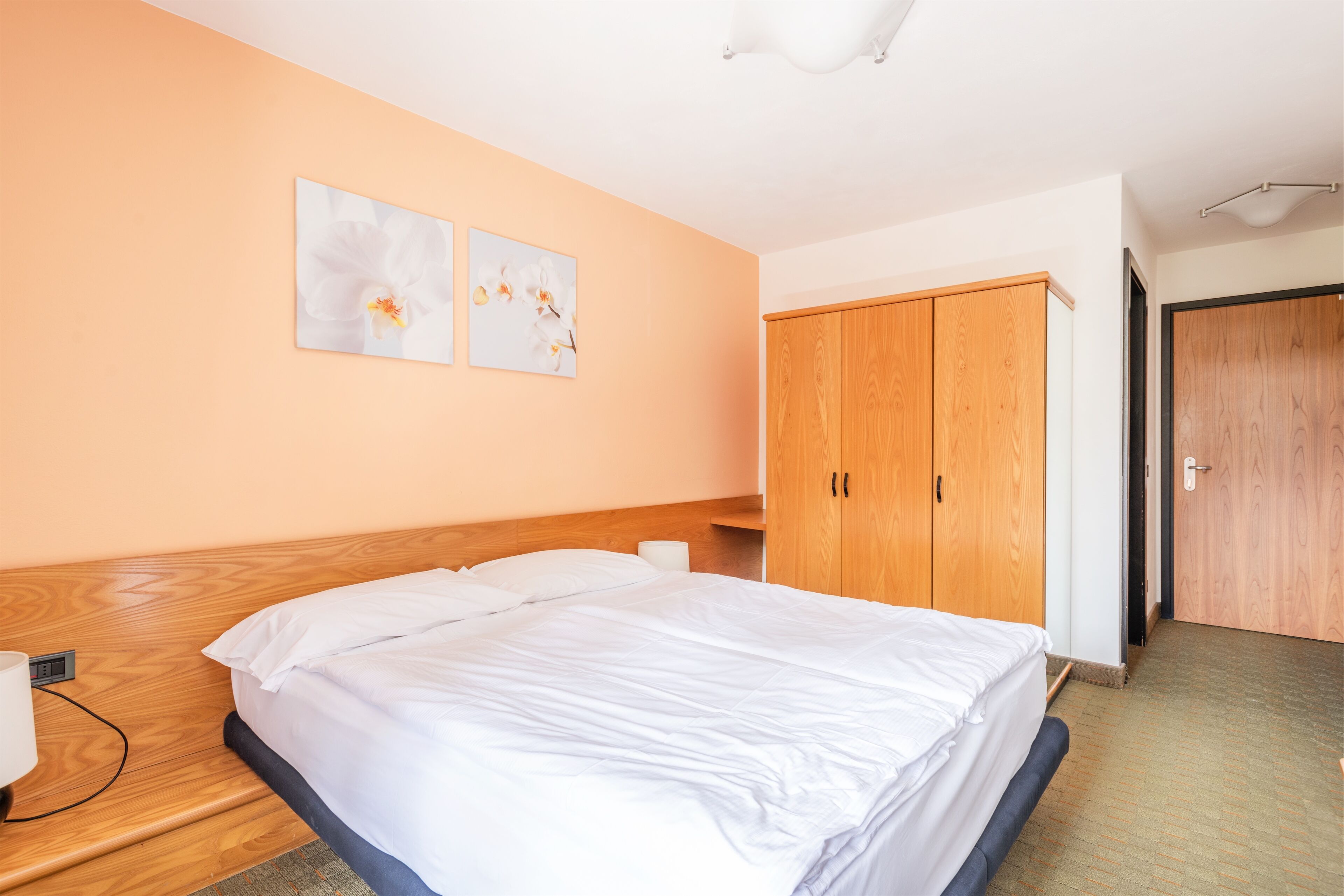 A hotel room with a bed, wooden wardrobe, and two wall-mounted flower paintings.