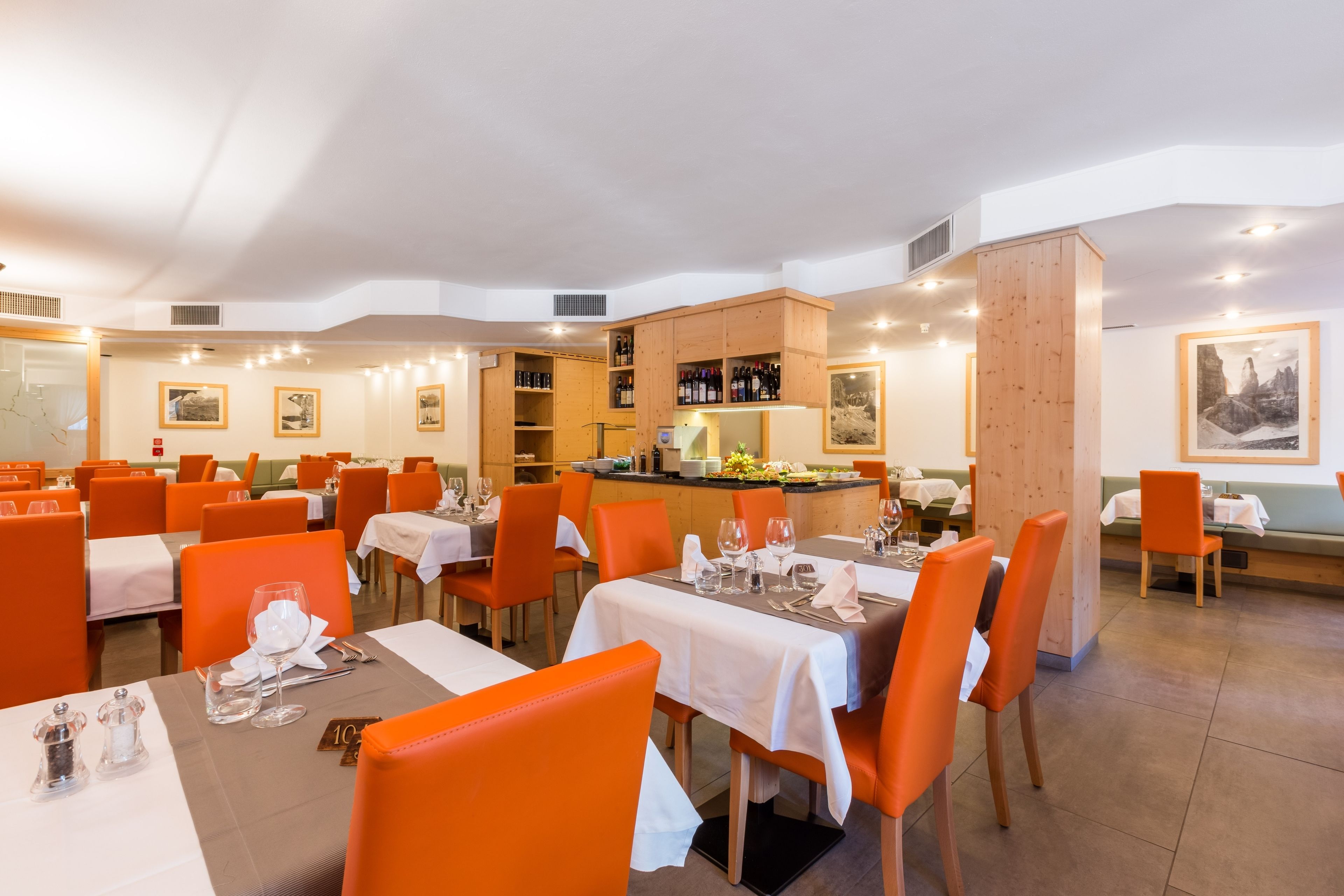 Un restaurante con varias mesas y sillas anaranjadas, una zona de cocina y cuadros en las paredes.