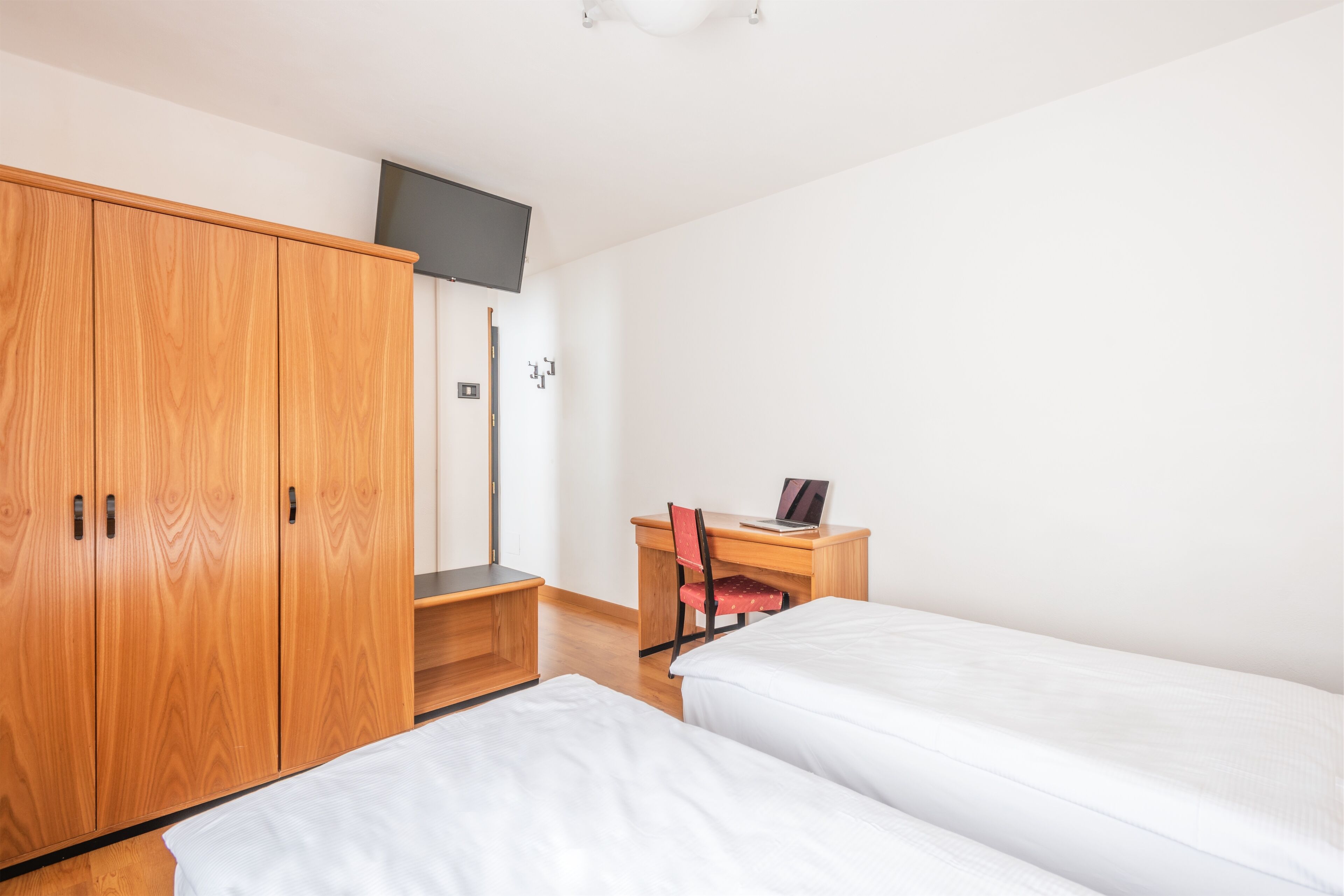 Una habitación con dos camas, un armario de madera, un escritorio con silla y un televisor fijado en la pared.