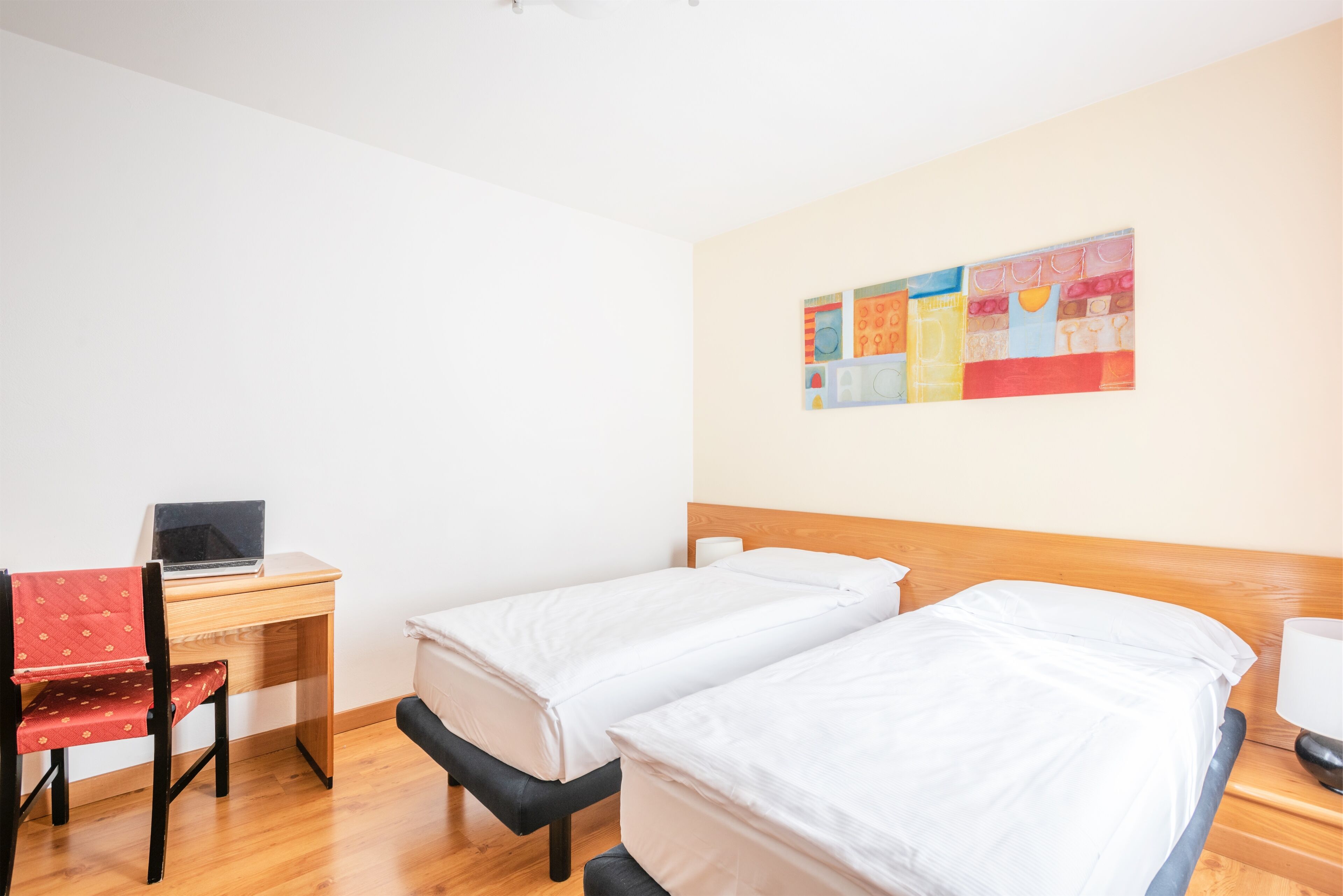 Una habitación con dos camas individuales, un escritorio con una computadora portátil, una silla y un colorido adorno en la pared.