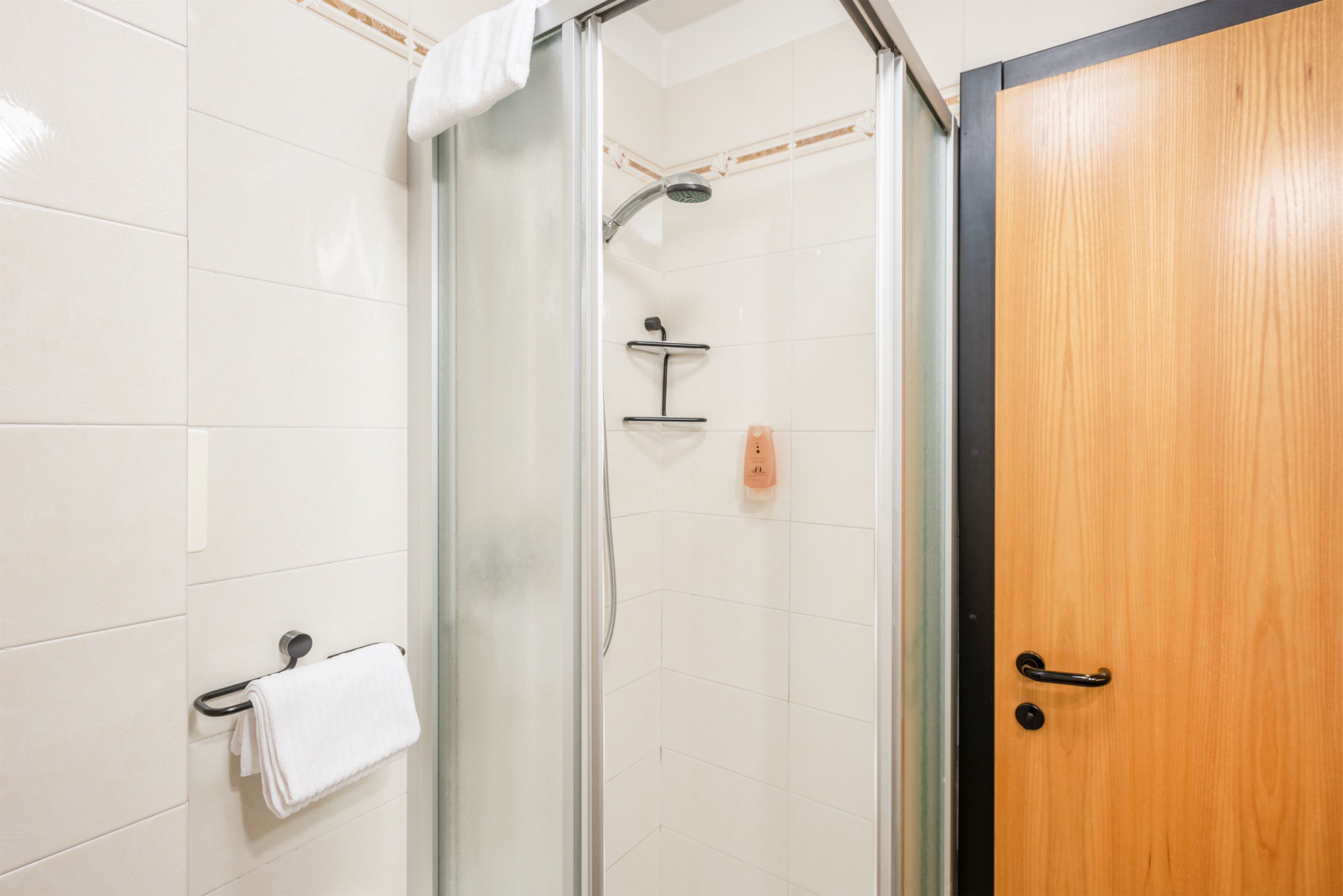 Una ducha con cerramiento de vidrio, un cabezal de ducha manual, un perchero y una puerta de madera con manija negra.