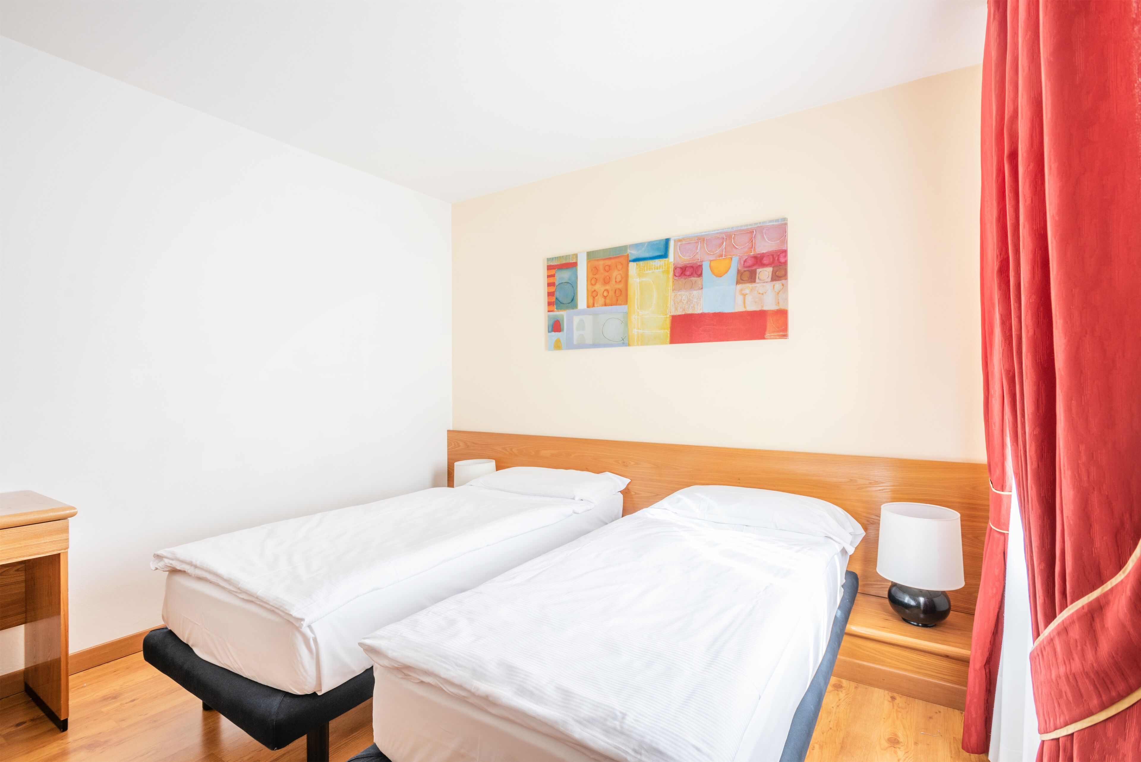 A hotel room with two single beds, a wooden headboard, a bedside table with a lamp, and a colorful abstract painting on the wall.