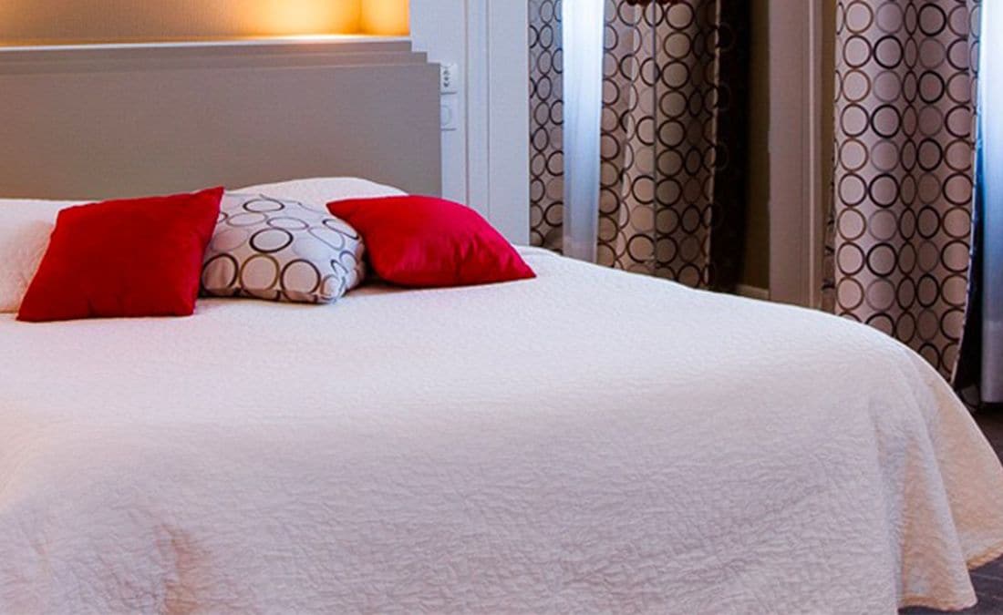 A neatly made bed with a white duvet and red pillows, a headboard, and patterned curtains.