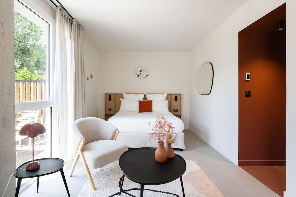 A modern bedroom with a bed, a chair, a small table, and a vase of dried flowers.