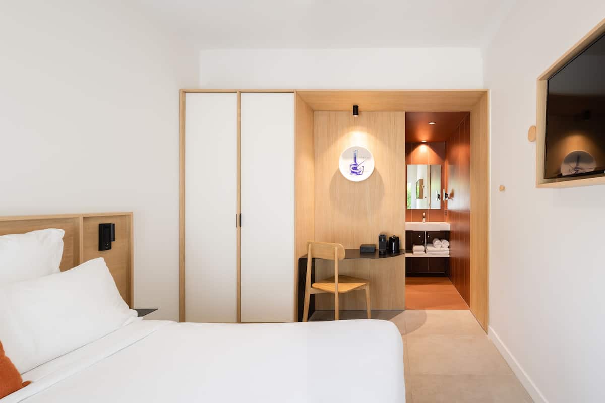 A hotel room with a bed, a desk, a chair, a wardrobe, and a bathroom with a sink and mirror.