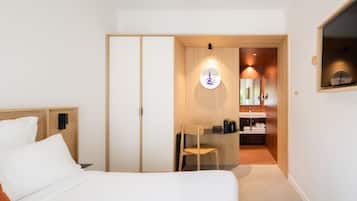 A hotel room with a bed, a desk, a chair, a wardrobe, and a bathroom with a sink and mirror.