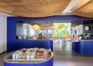 A modern breakfast buffet with a variety of food items and beverages, a large window with a view of greenery, and a ceiling with a woven texture. - Les Elmes - Hôtel, Spa & Plage Privée (Banyuls-sur-Mer)