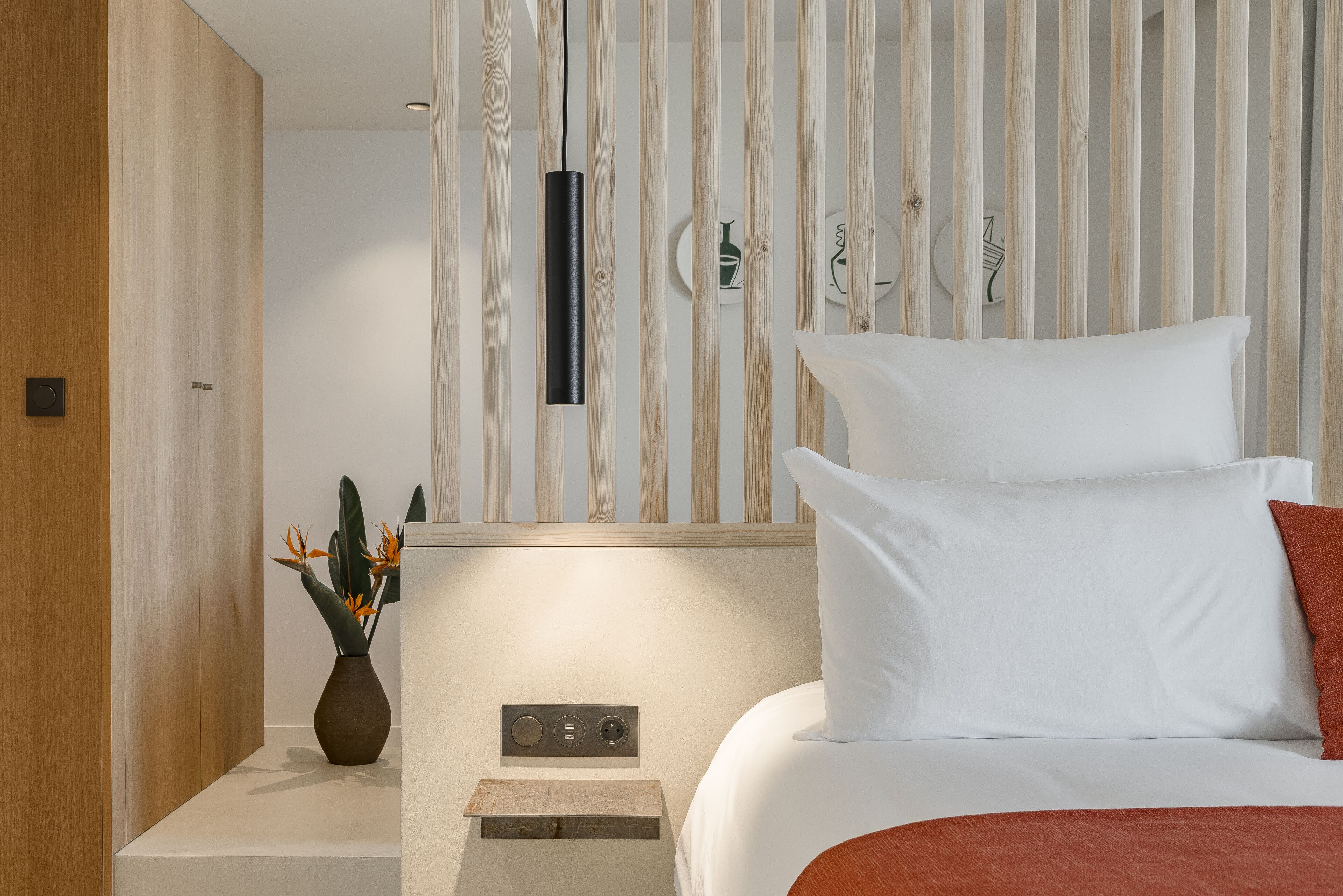 Un dormitorio moderno con un armario de madera, una mesita de noche con un florero y una cama con ropa de cama blanca y roja, además de una lámpara de pared.