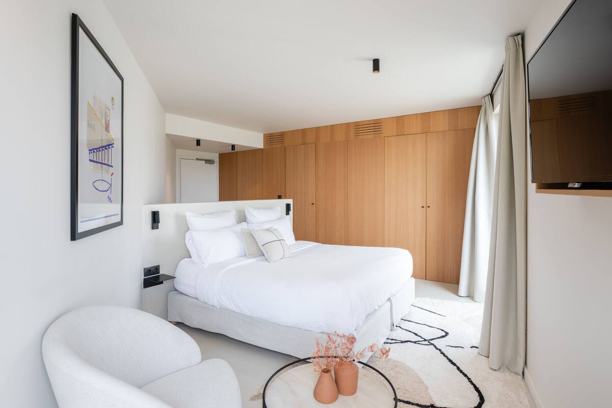 A modern bedroom with a large bed, a white armchair, and a wooden wardrobe.