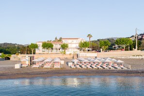 A beachside hotel with multiple lounge chairs and umbrellas, a building with a sign, and a view of the sea. - Les Elmes - Hôtel, Spa & Plage Privée (Banyuls-sur-Mer)
