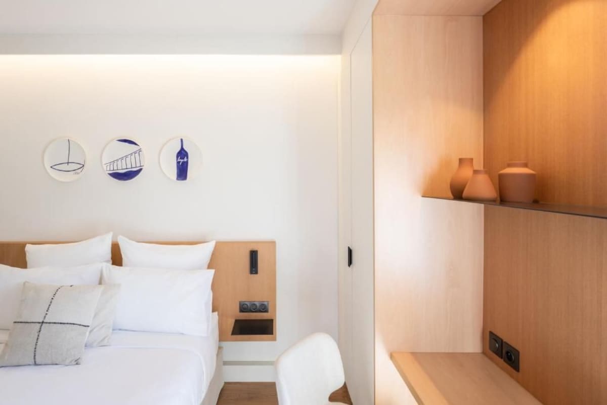 Una habitación de hotel moderna con una cama, un cabecero de madera, un pequeño estante con jarrones y una obra de arte en la pared que representa una botella y un puente.