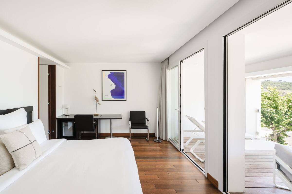 A hotel room with a bed, a desk, a chair, and a large window with a view of greenery.