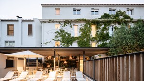 Un área de piscina en la azotea con sillones de descanso, una zona de estar cubierta y vistas a un edificio blanco con ventanas y vegetación.