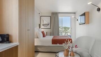 A modern hotel room with a bed, a round glass table with vases, a white armchair, and a large window with a view of greenery.