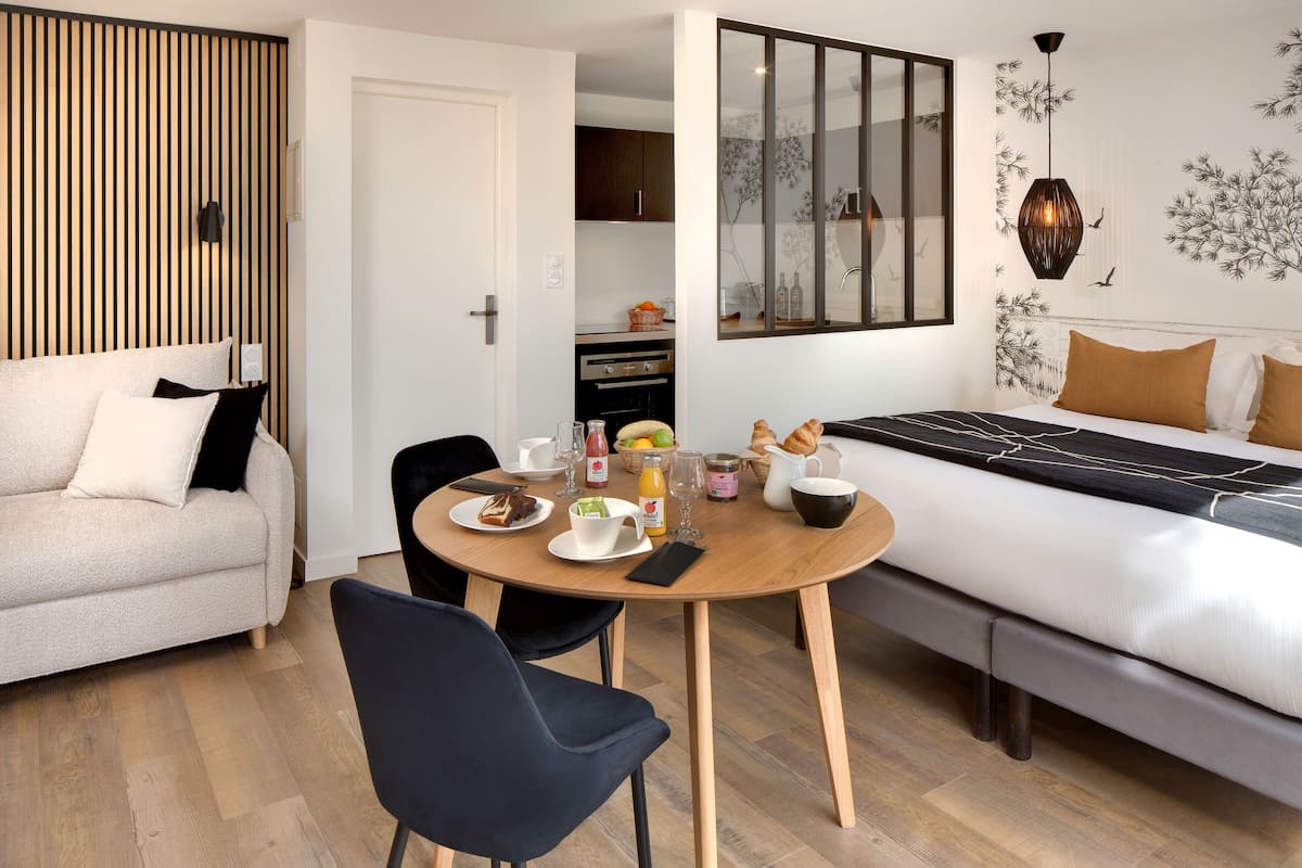 A modern bedroom with a bed, a dining table with breakfast items, a sofa, and a kitchen area.