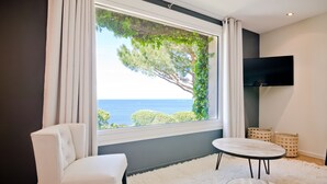 Habitación moderna con un amplio ventanal que ofrece vistas al mar y a los árboles, un sofá blanco, una mesa redonda y un televisor de pantalla plana.