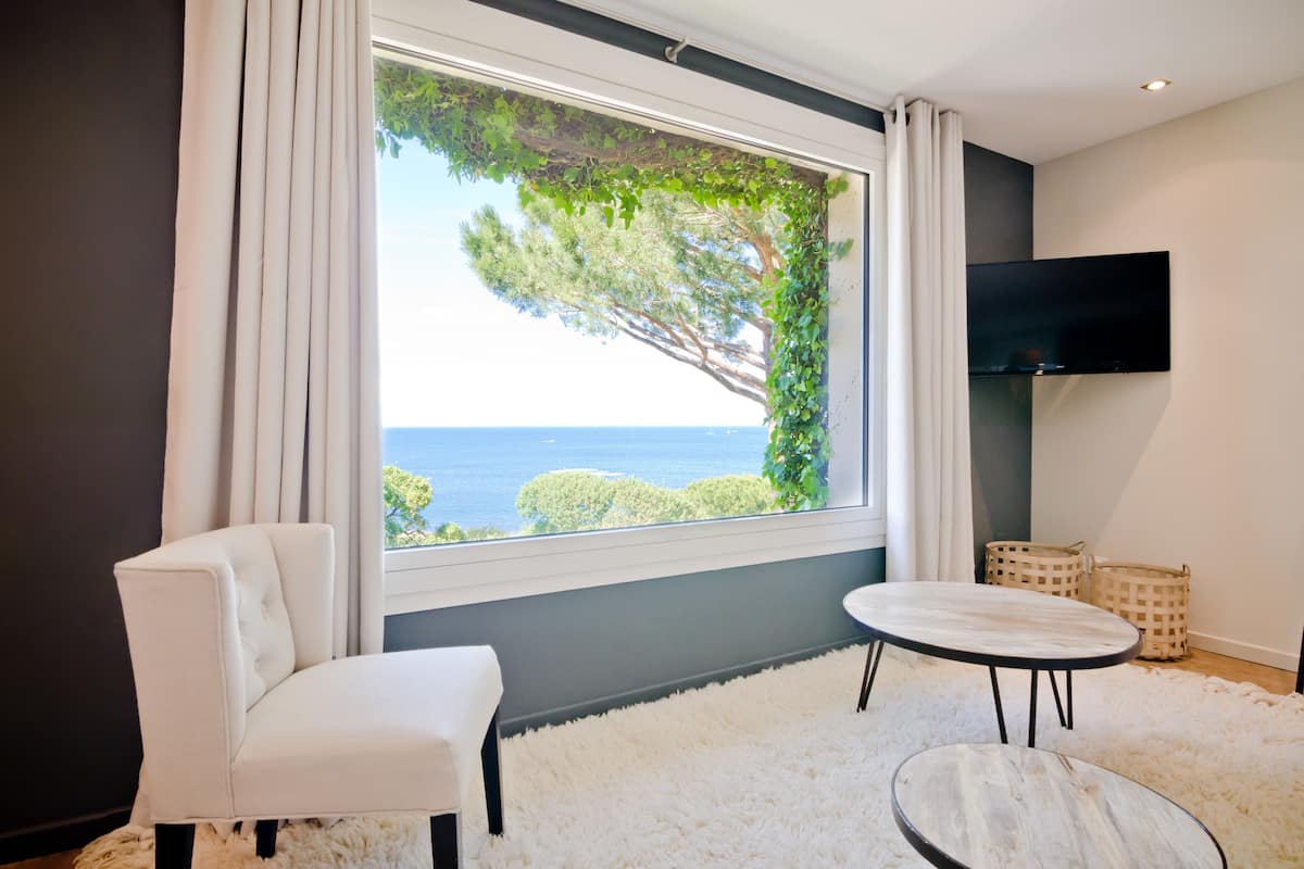 A modern room with a large window offering a view of the sea and trees, a white sofa, a round table, and a mounted TV.