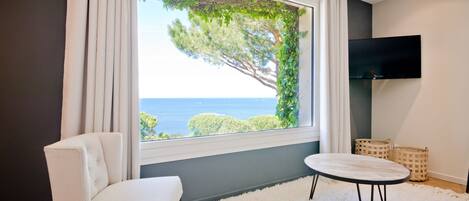 Habitación moderna con un amplio ventanal que ofrece vistas al mar y a los árboles, un sofá blanco, una mesa redonda y un televisor de pantalla plana.