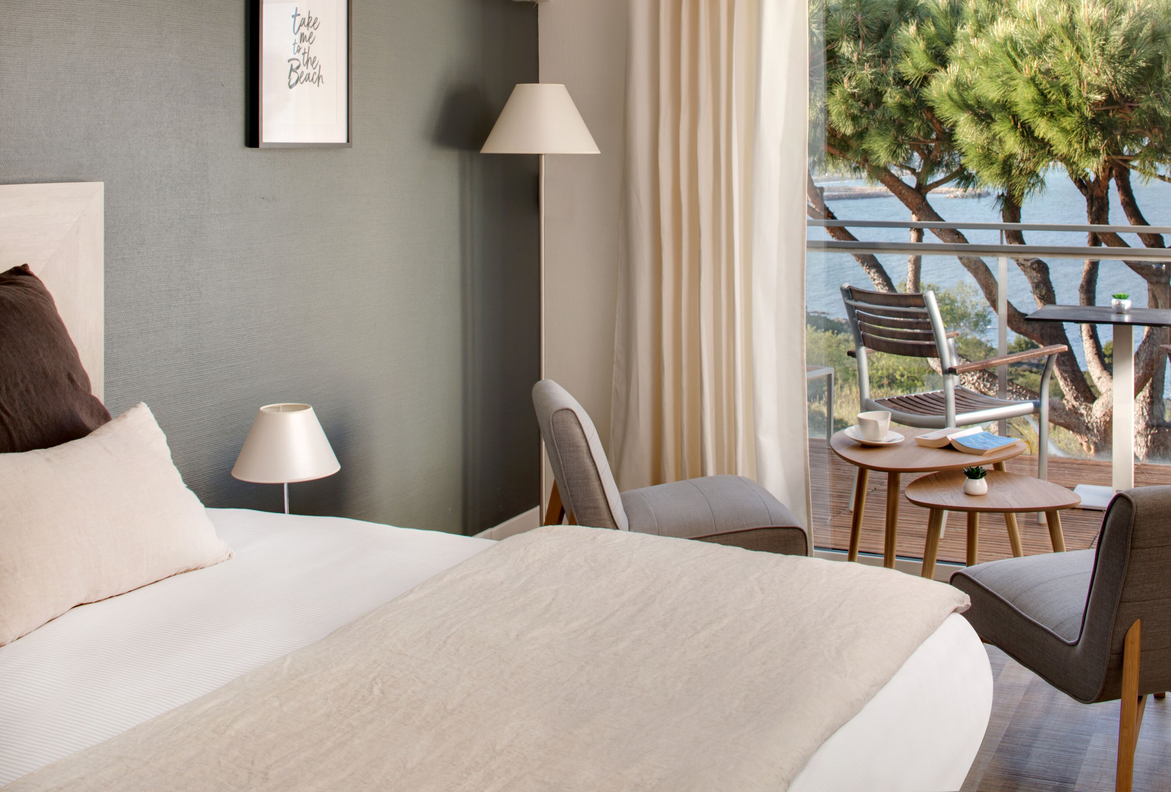 A hotel room with a large bed, a desk with a chair, a lamp, and a view of the sea.