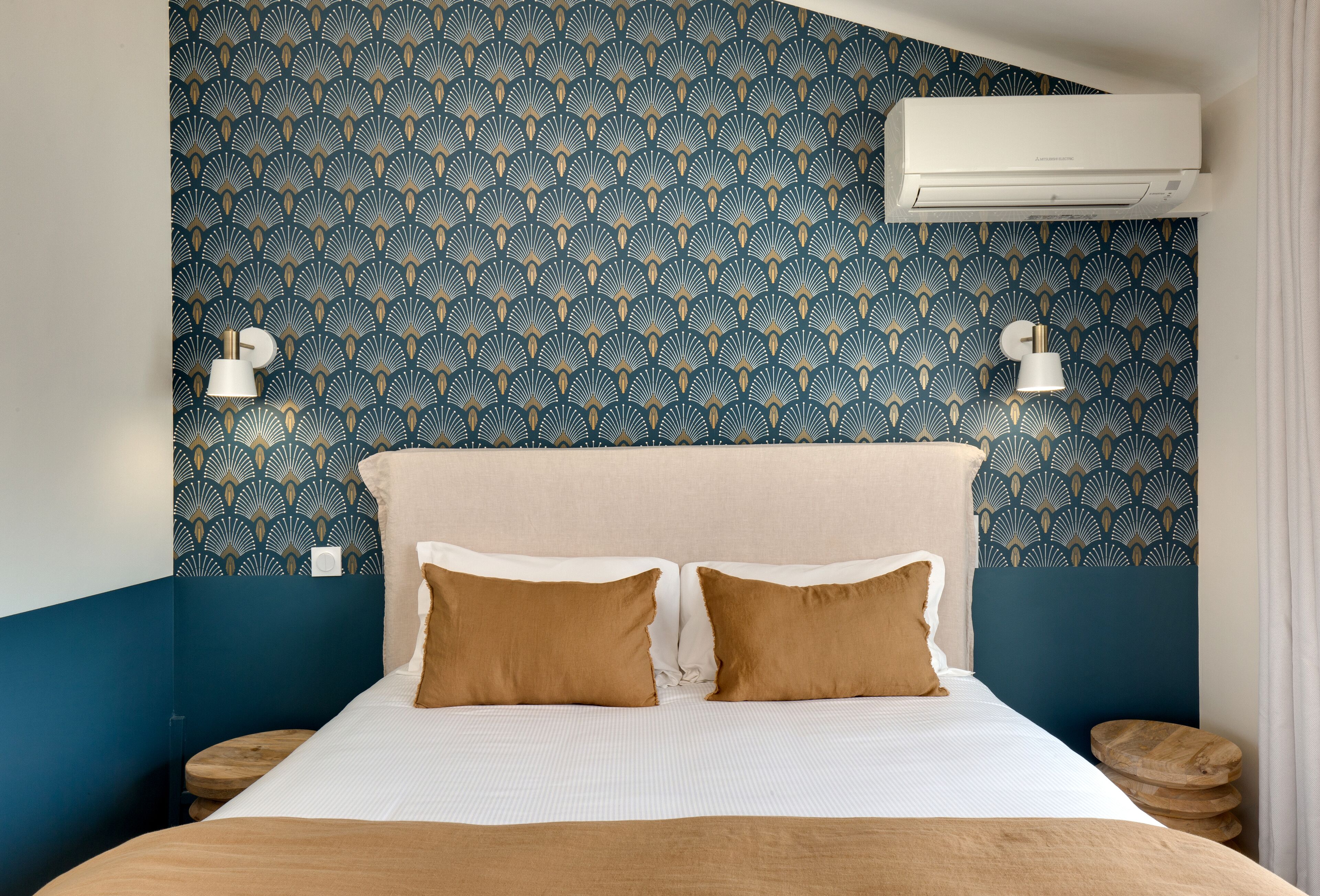 A hotel room with a patterned wallpaper, a bed with two pillows, bedside tables, and an air conditioning unit.