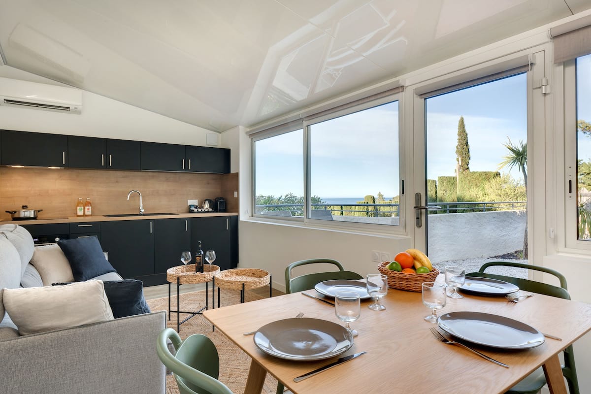 A modern kitchen with dark cabinets and a wooden countertop, a dining area with a table set for two, and a large window offering a view of the outdoors.