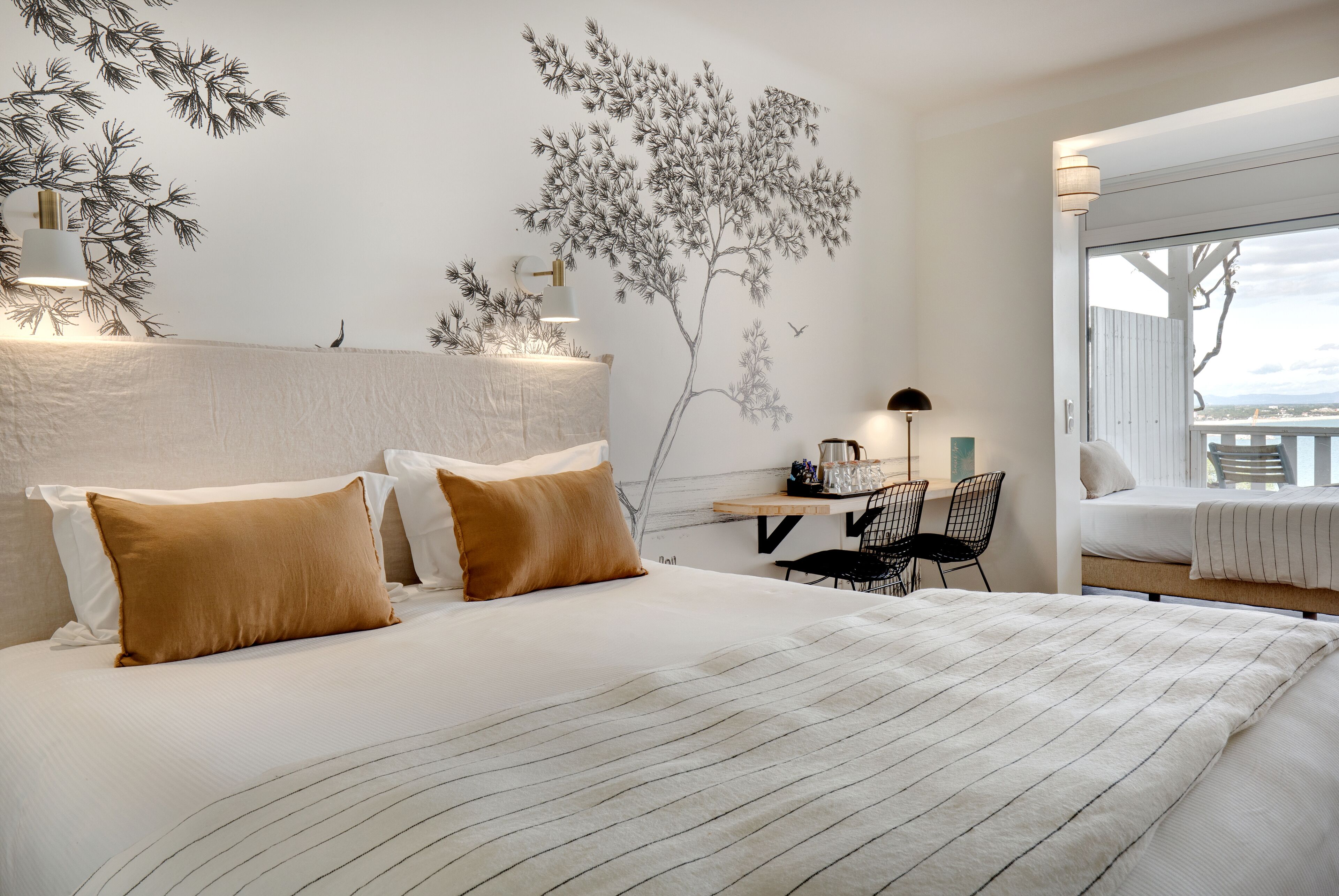 Une chambre d’hôtel avec un grand lit, deux oreillers, un bureau avec une chaise, une fresque murale et une fenêtre donnant sur un paysage.