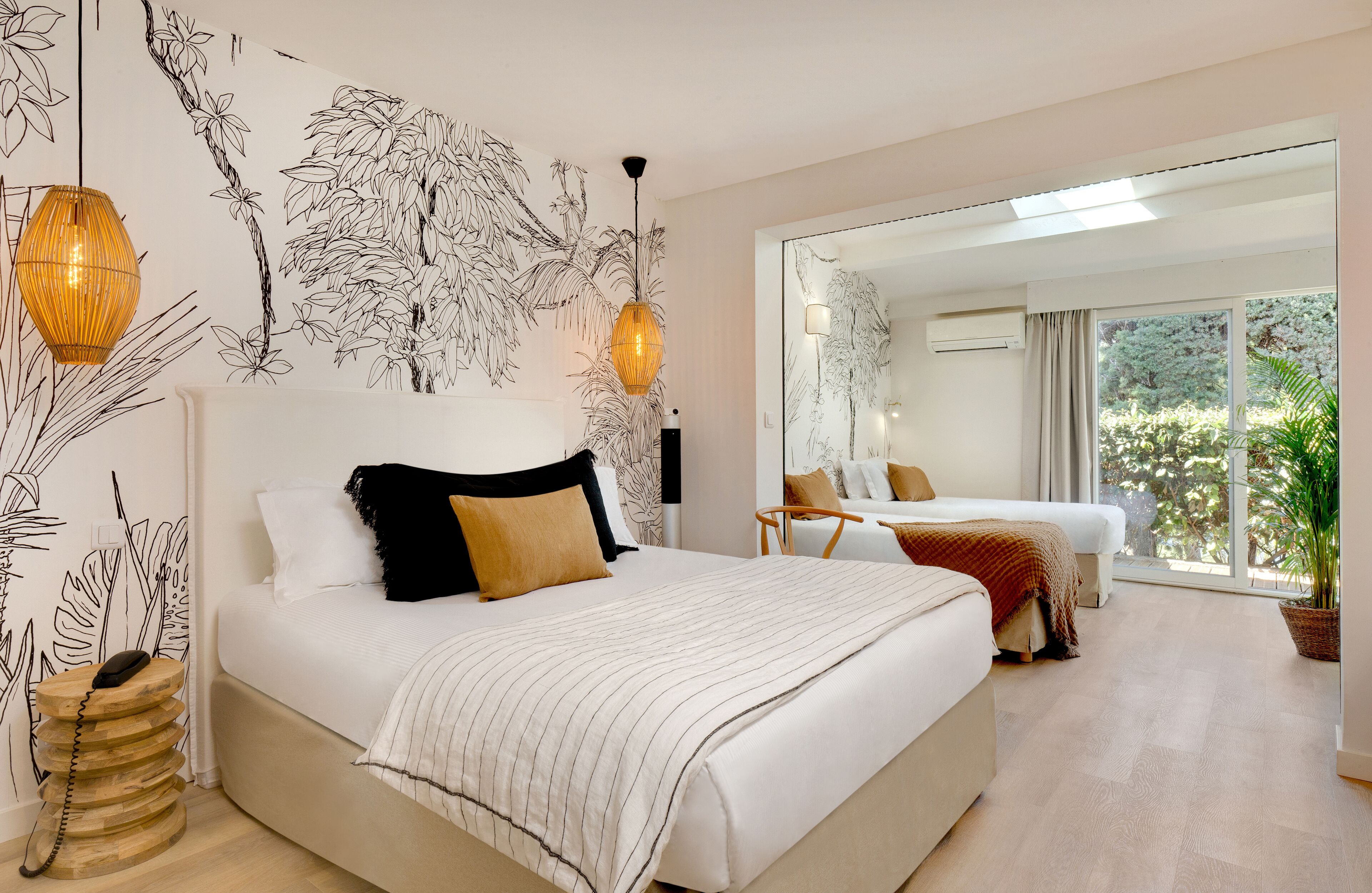 Un dormitorio moderno con una cama grande, una cama más pequeña, un sofá, una lámpara colgante y un amplio ventanal con vistas a la vegetación.