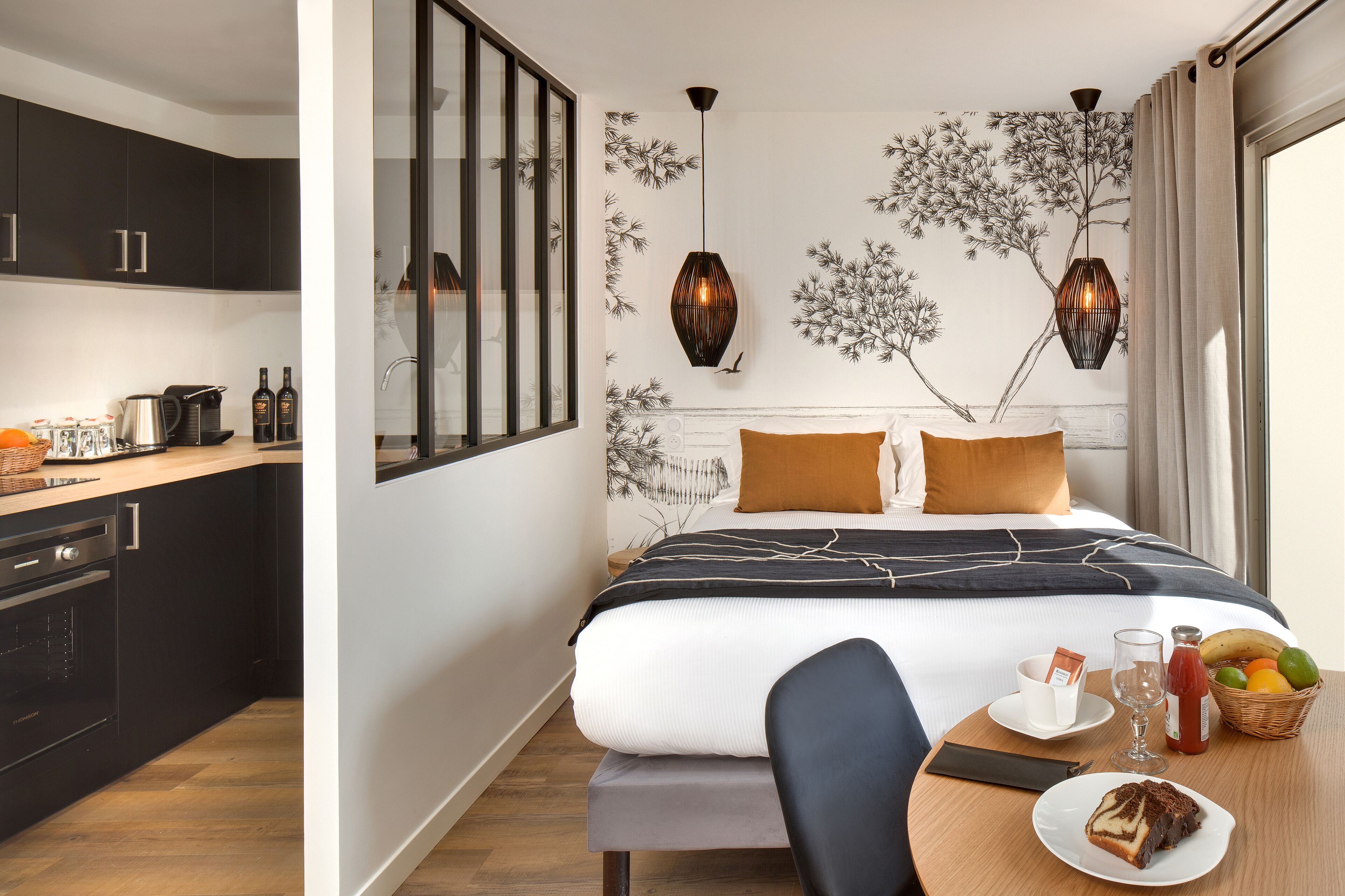 Quarto de hotel moderno com cozinha, cama com colcha listrada em preto e branco, bandeja de café da manhã com uma fatia de bolo e uma fruteira.