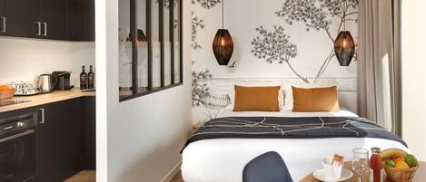 Quarto de hotel moderno com cozinha, cama com colcha listrada em preto e branco, bandeja de café da manhã com uma fatia de bolo e uma fruteira.