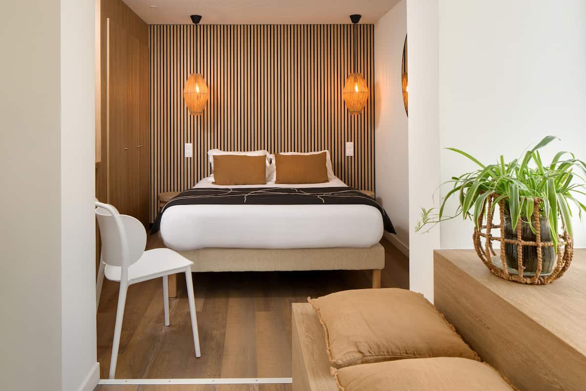A modern bedroom with a bed, two bedside lamps, a white chair, a wooden floor, and a potted plant.