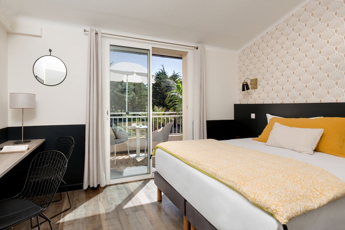 A hotel room with a bed, a desk, a chair, a mirror, and a balcony with a table and chairs.