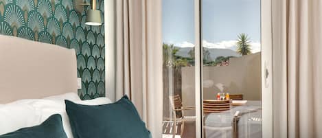 Una habitación de hotel con una cama, almohadones color turquesa, una pared con diseño, una ventana que da a un balcón y una mesa con sillas.