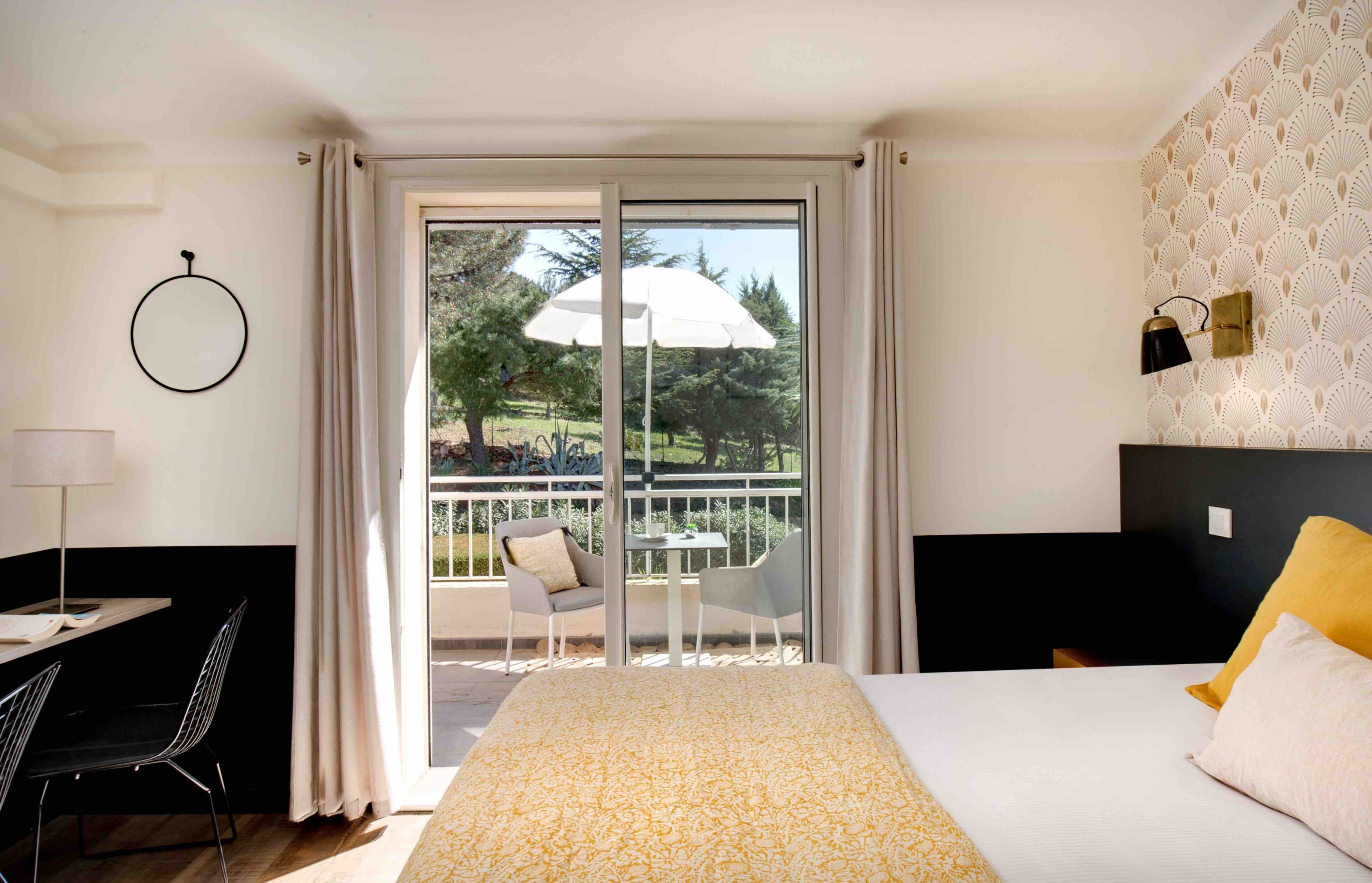 A hotel room with a bed, a desk, a chair, a balcony with a table and chairs, and a view of trees.