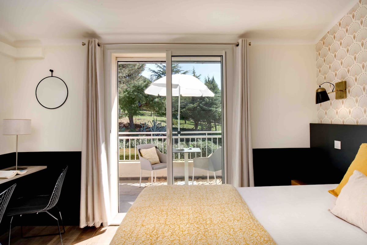 A hotel room with a bed, a desk, a chair, a balcony with a table and chairs, and a view of trees.