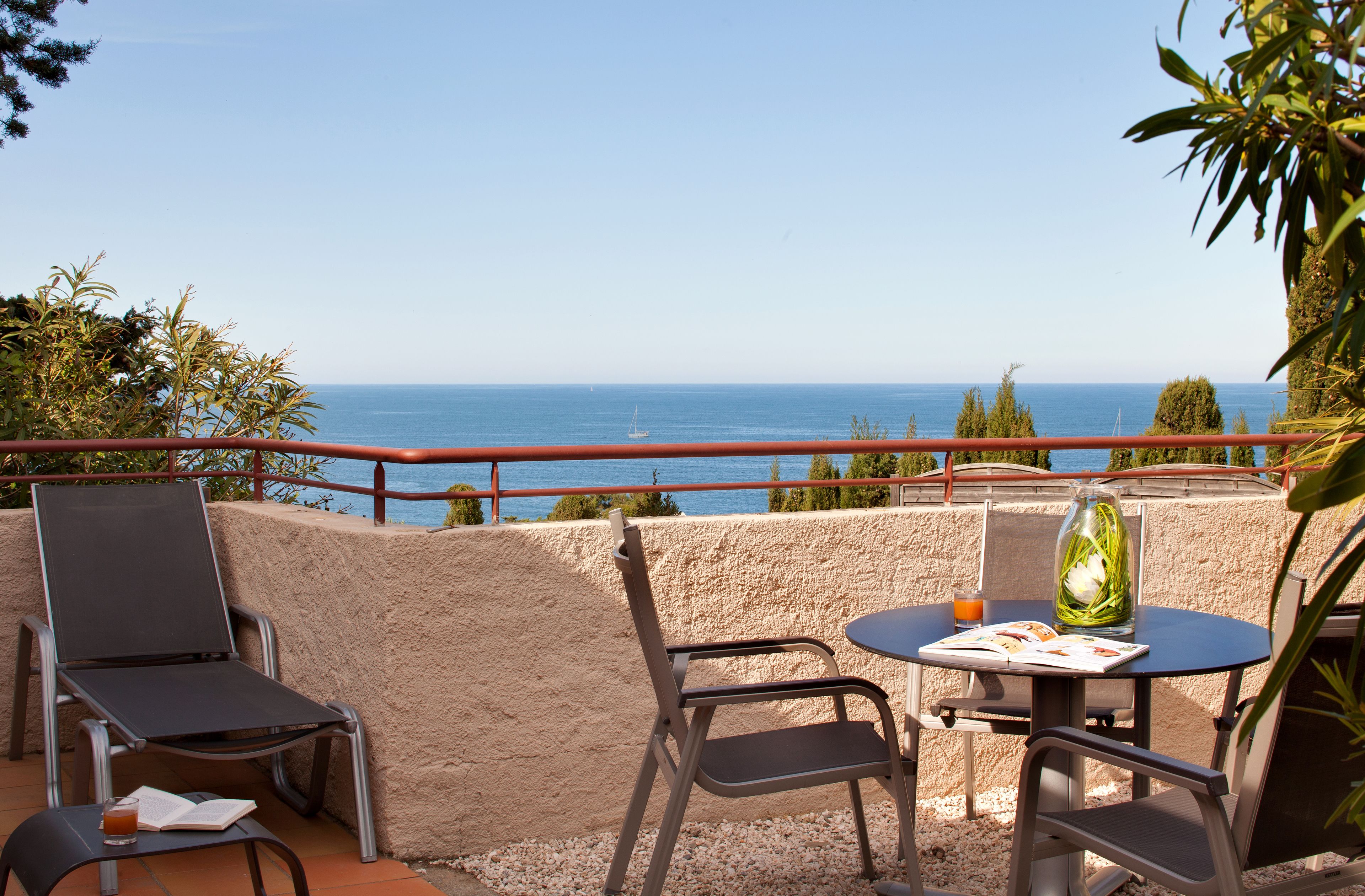 A terrace with a table and chairs, a book, a glass of drink, and a pitcher of flowers.